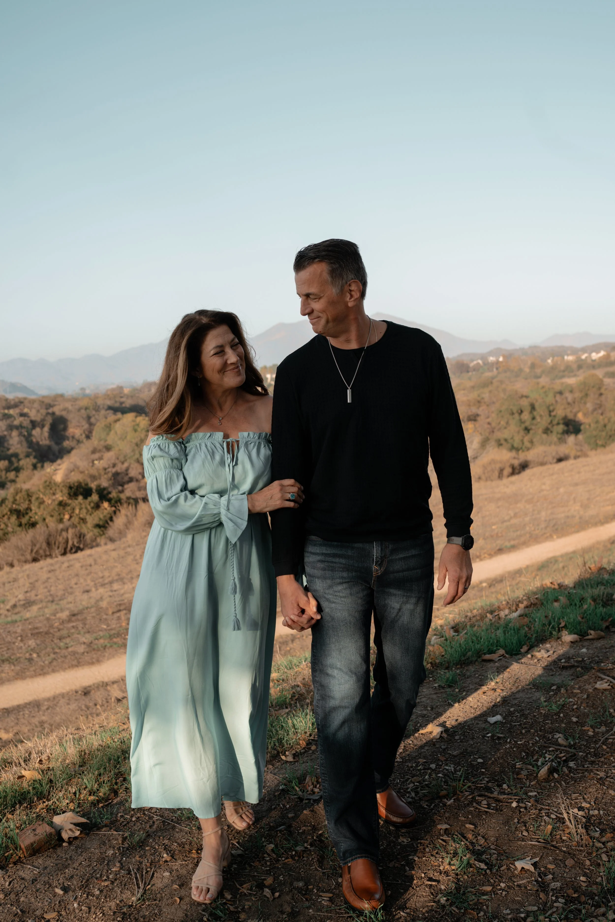 A man and woman walking hand in hand outdoors on a dirt path, smiling at each other with mountains and a clear sky in the background.