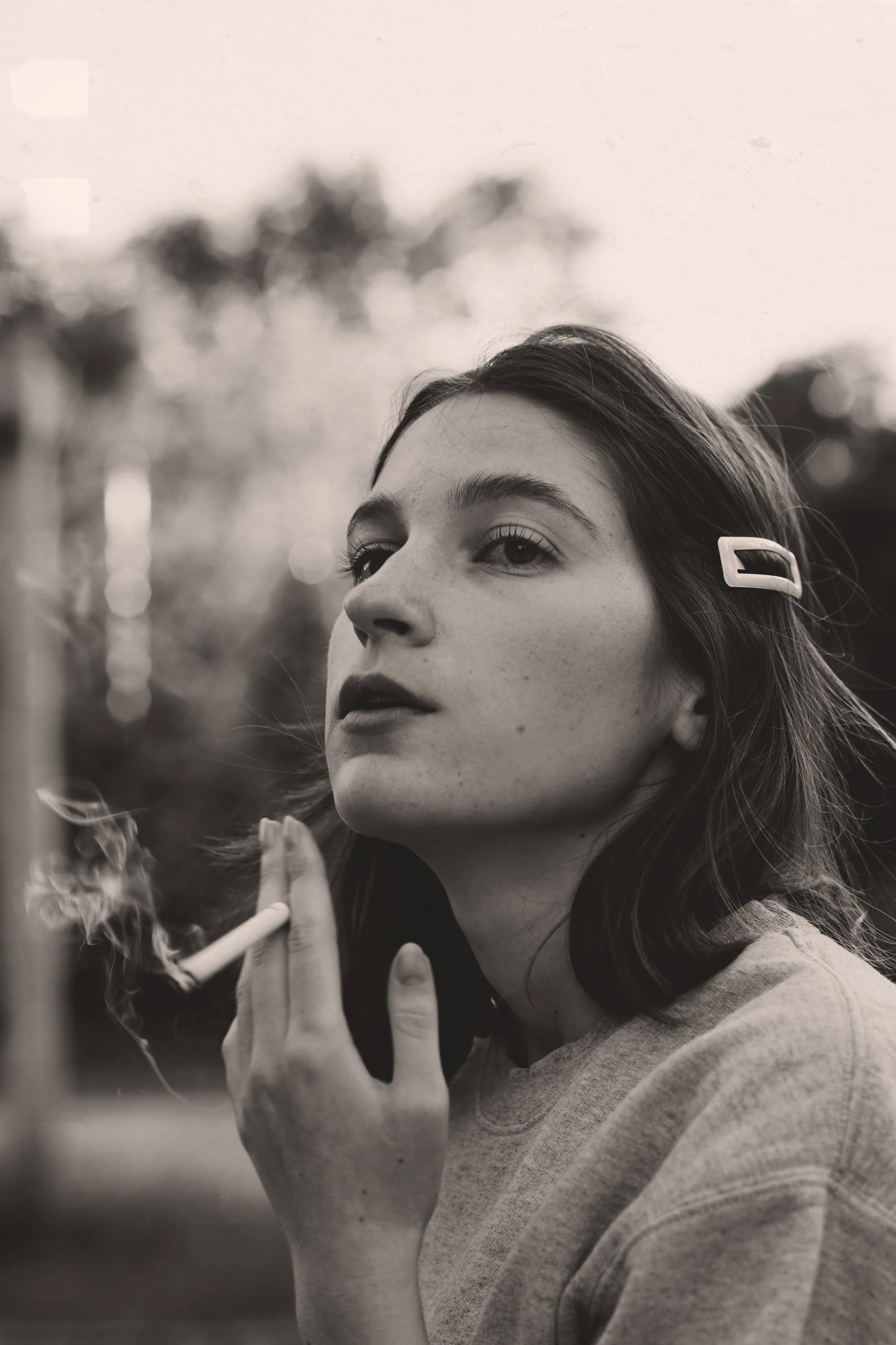 A young woman with dark hair and a hair clip, holding a cigarette, appears to be lost in thought outdoors.