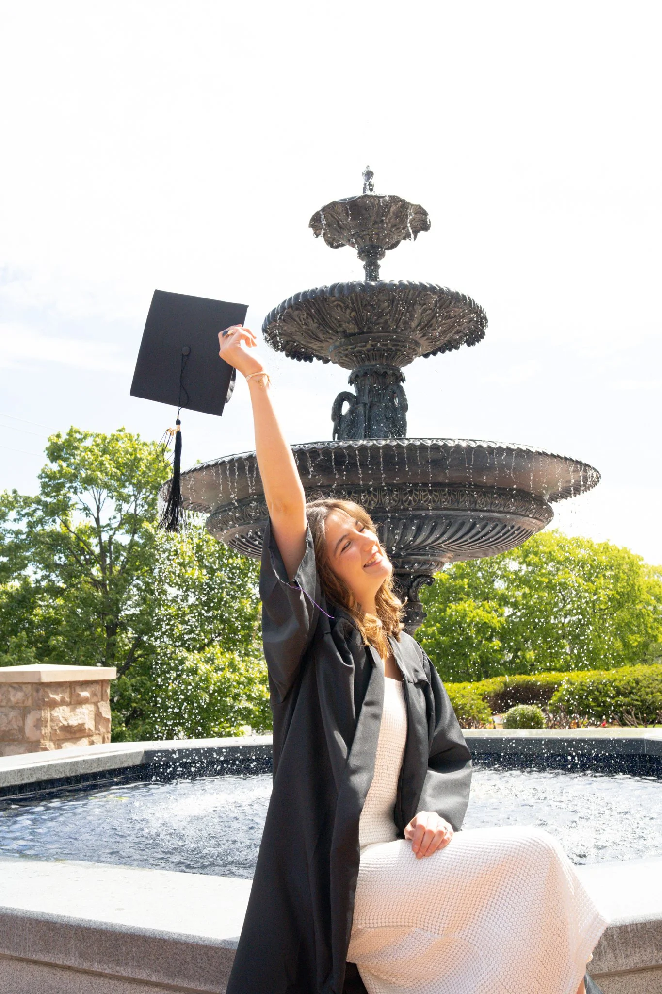 A young woman in graduation gown holding her cap in one hand and smiling while sitting on the edge of a fountain with a multi-tiered decorative water fountain in the background, surrounded by trees and greenery on a sunny day.
