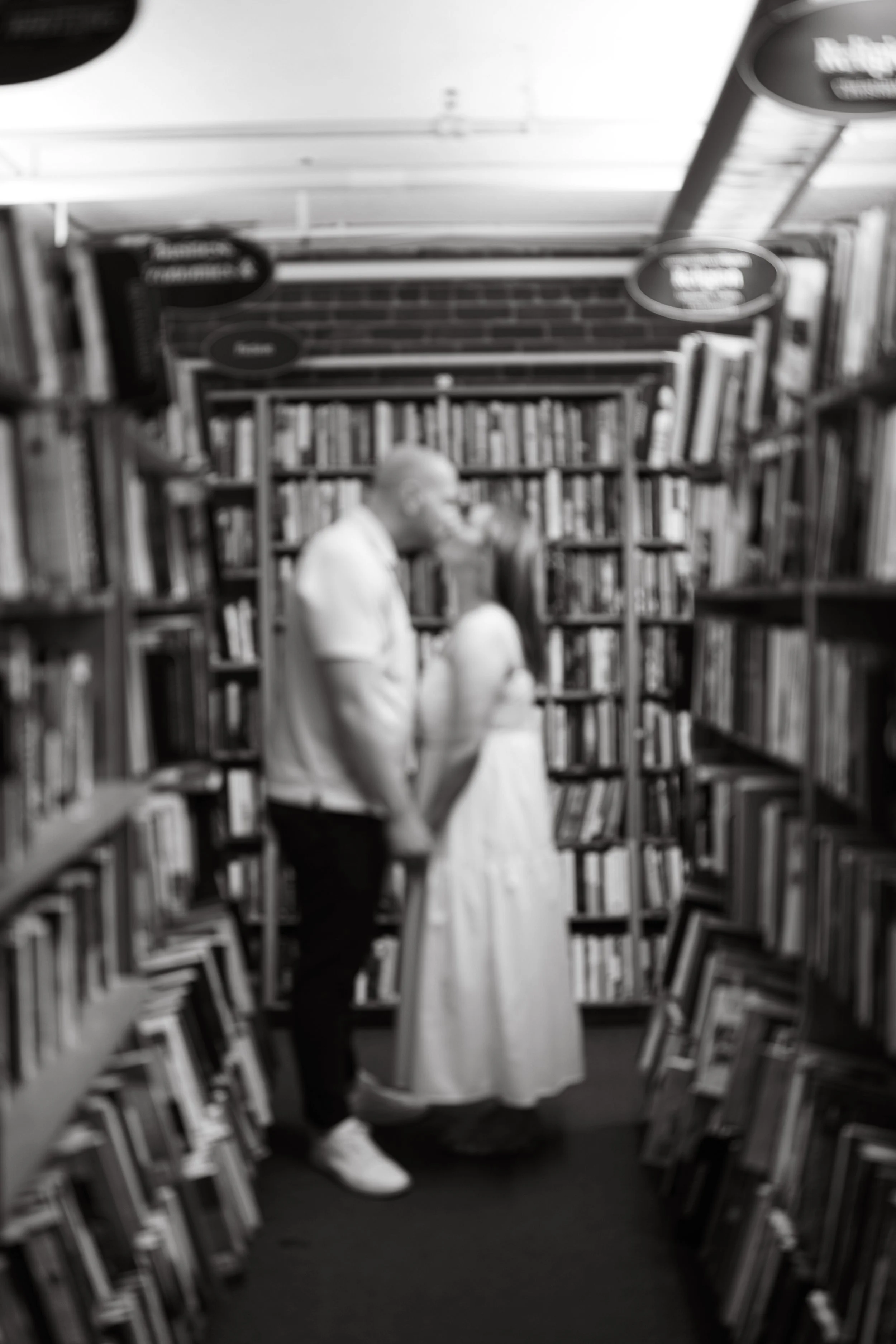 Two people, a man and a woman, holding hands and walking down an aisle in a library surrounded by tall bookshelves filled with books.