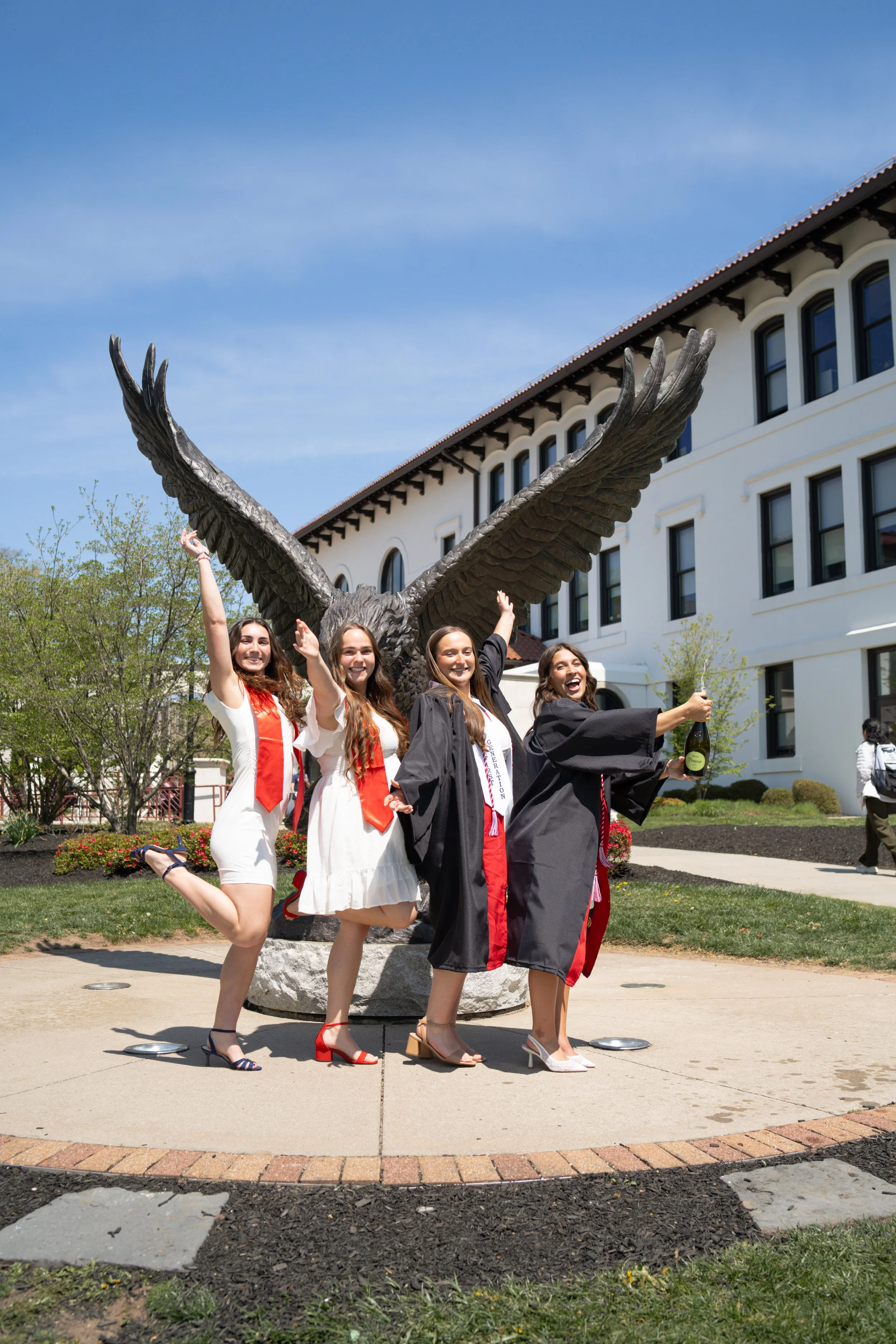 Four women in graduation gowns and caps celebrating in front of a large eagle statue outside a white building with multiple windows, during a sunny day.