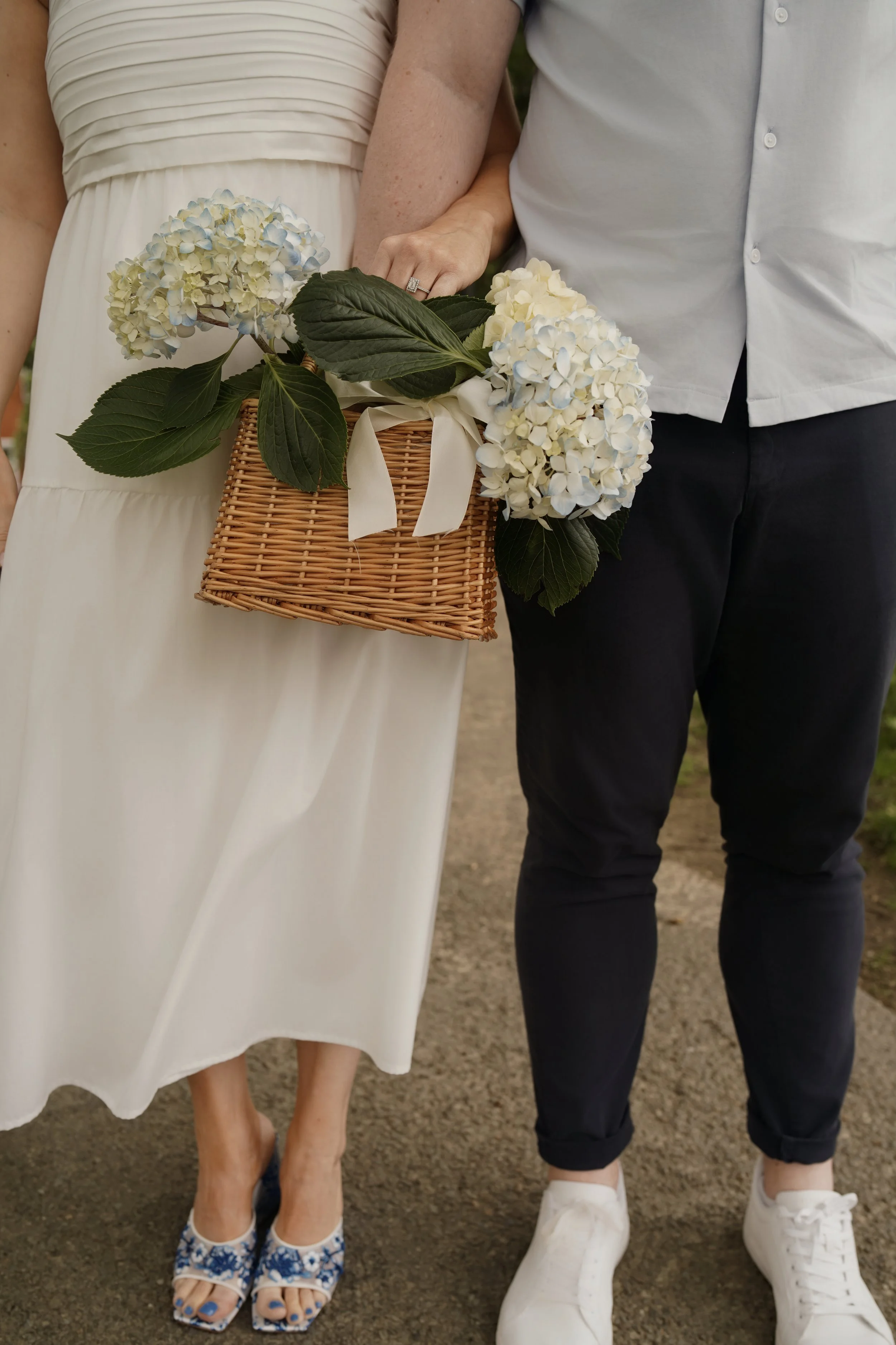 Close-up of a woman in a white dress holding a basket of blue and white hydrangeas, standing next to a man in black pants and white sneakers, both standing outdoors.