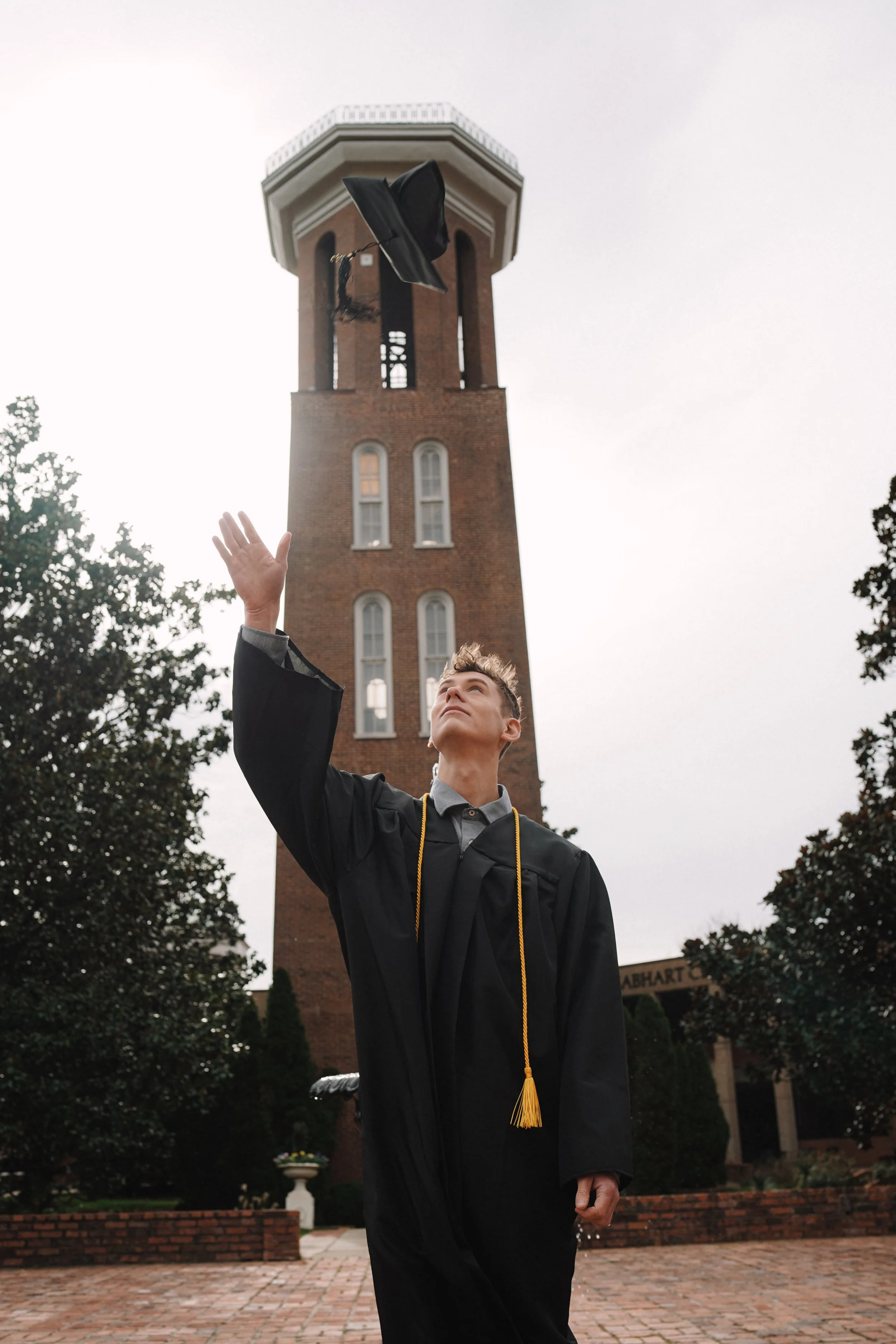 Young man in graduation cap and gown tossing cap in the air in front of a brick clock tower.
