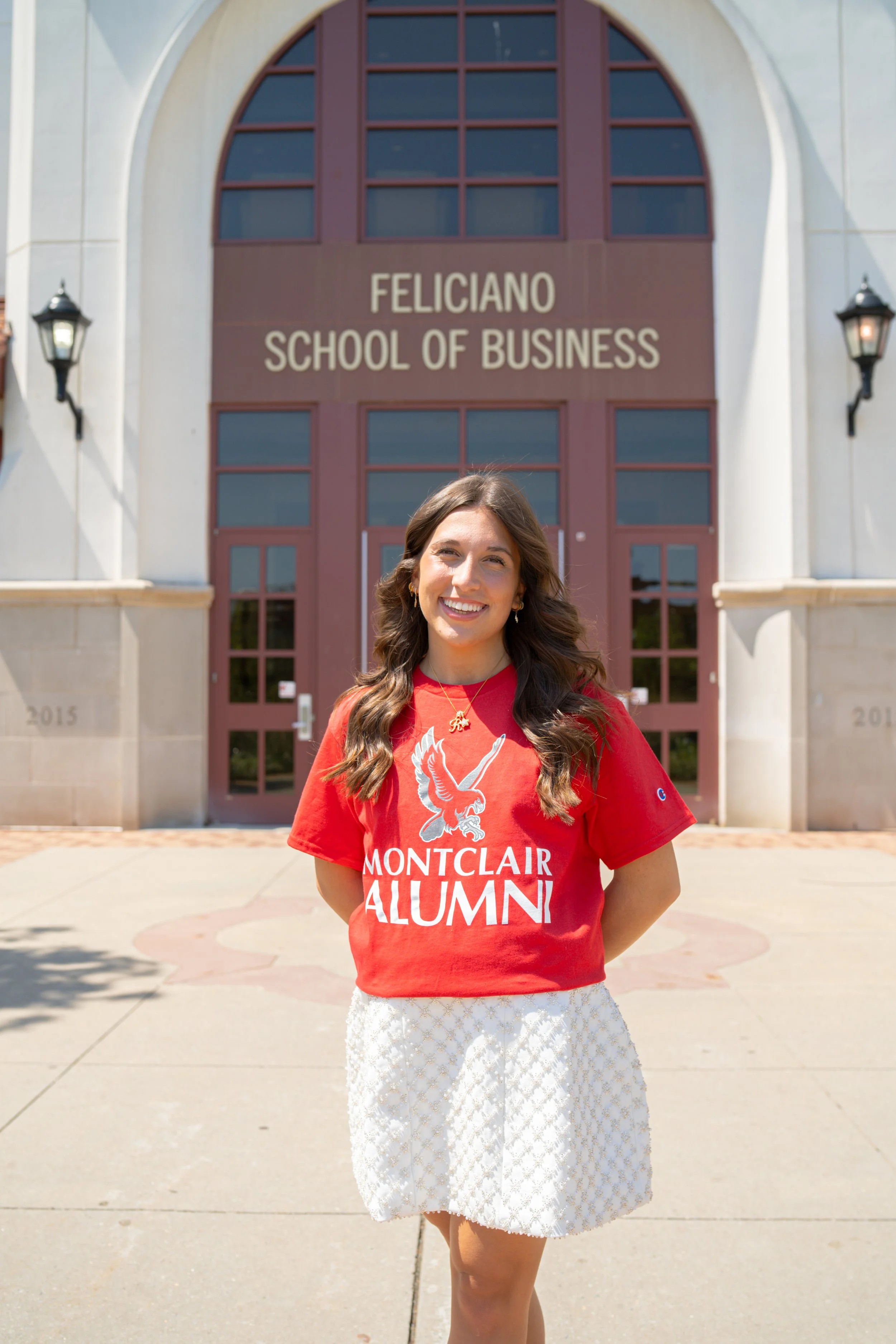 A young woman with long brown hair is standing outdoors in front of a large building with the sign 'Feliciano School of Business.' She is wearing a red T-shirt that says 'Montclair Alum' and a white skirt with a floral pattern. She is smiling and loo