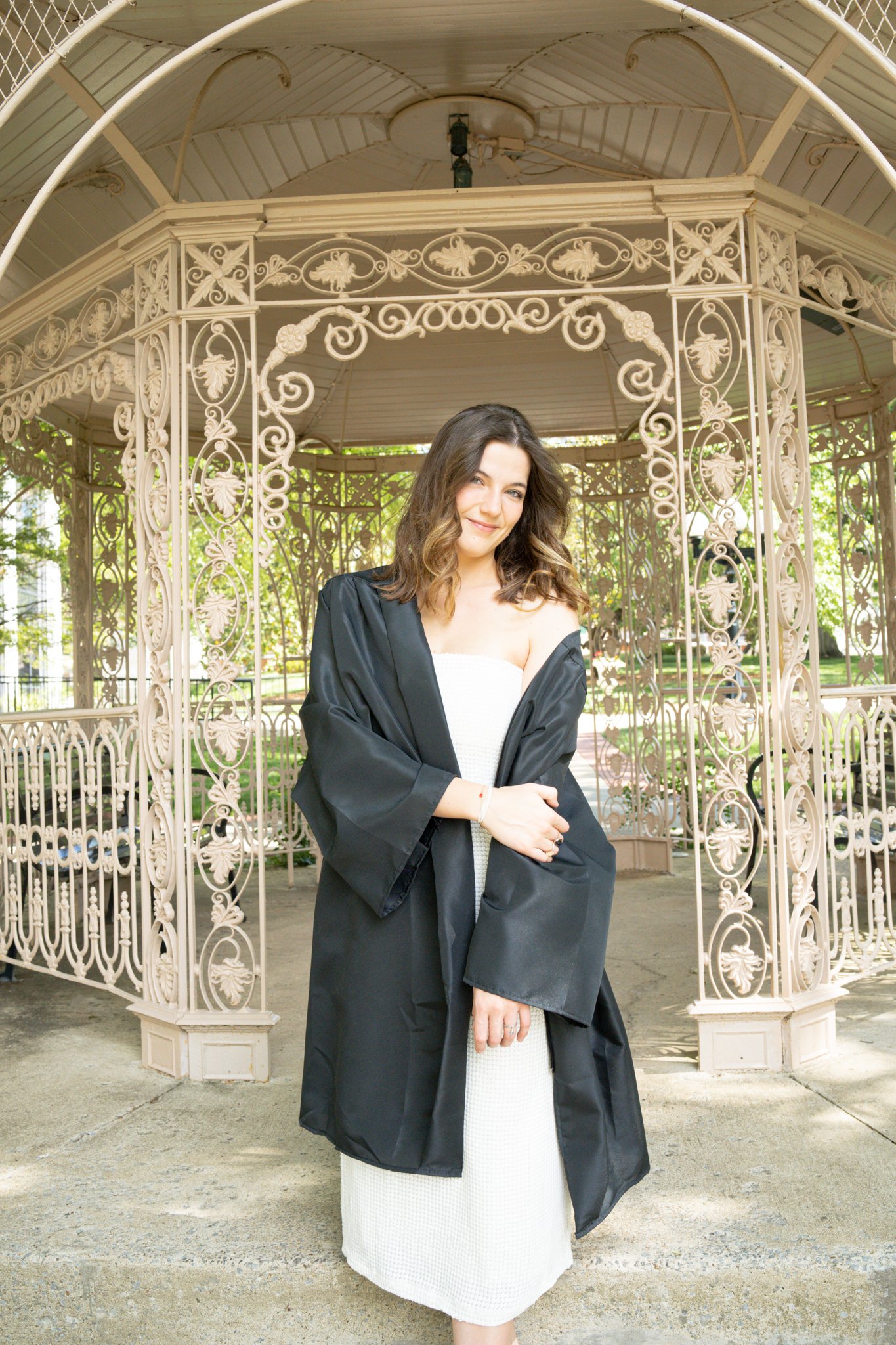A woman wearing a white dress with a black graduation gown, standing outdoors under a decorative gazebo with an intricate iron design.