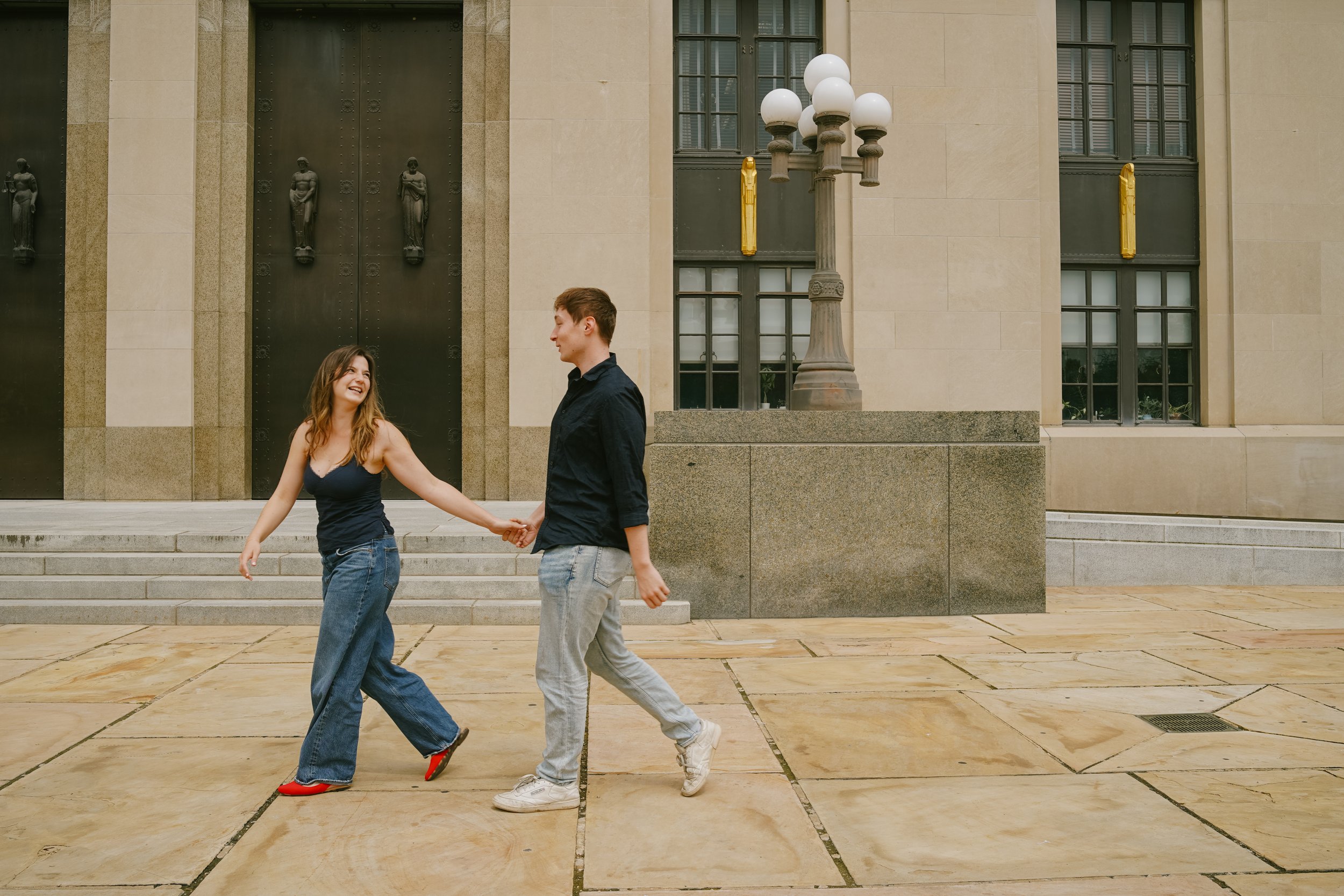 Two young adults, a woman with long brown hair wearing a black tank top and baggy jeans, and a man with short brown hair wearing a black shirt and light gray pants, are walking and holding hands outside near a large building with tall windows and bla