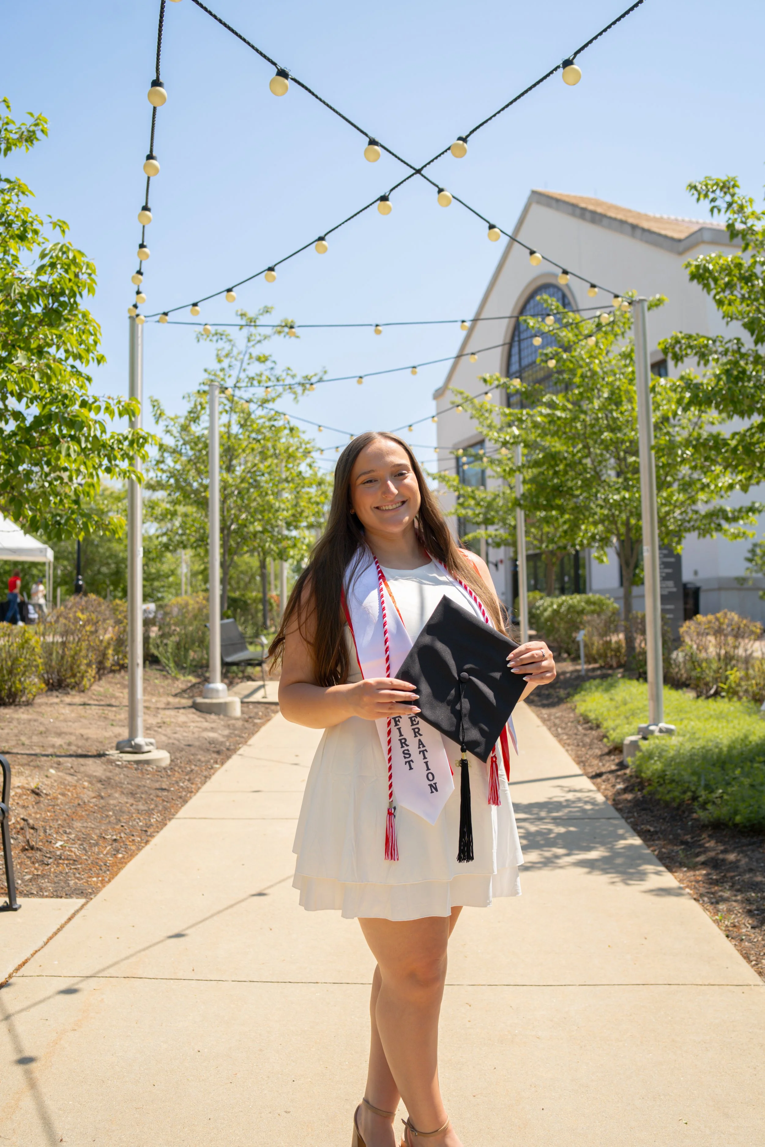 A young woman in a white dress holding a black graduation cap, smiling outdoors under string lights, with trees and a church in the background, celebrating her graduation with a sash that reads "First Generation."