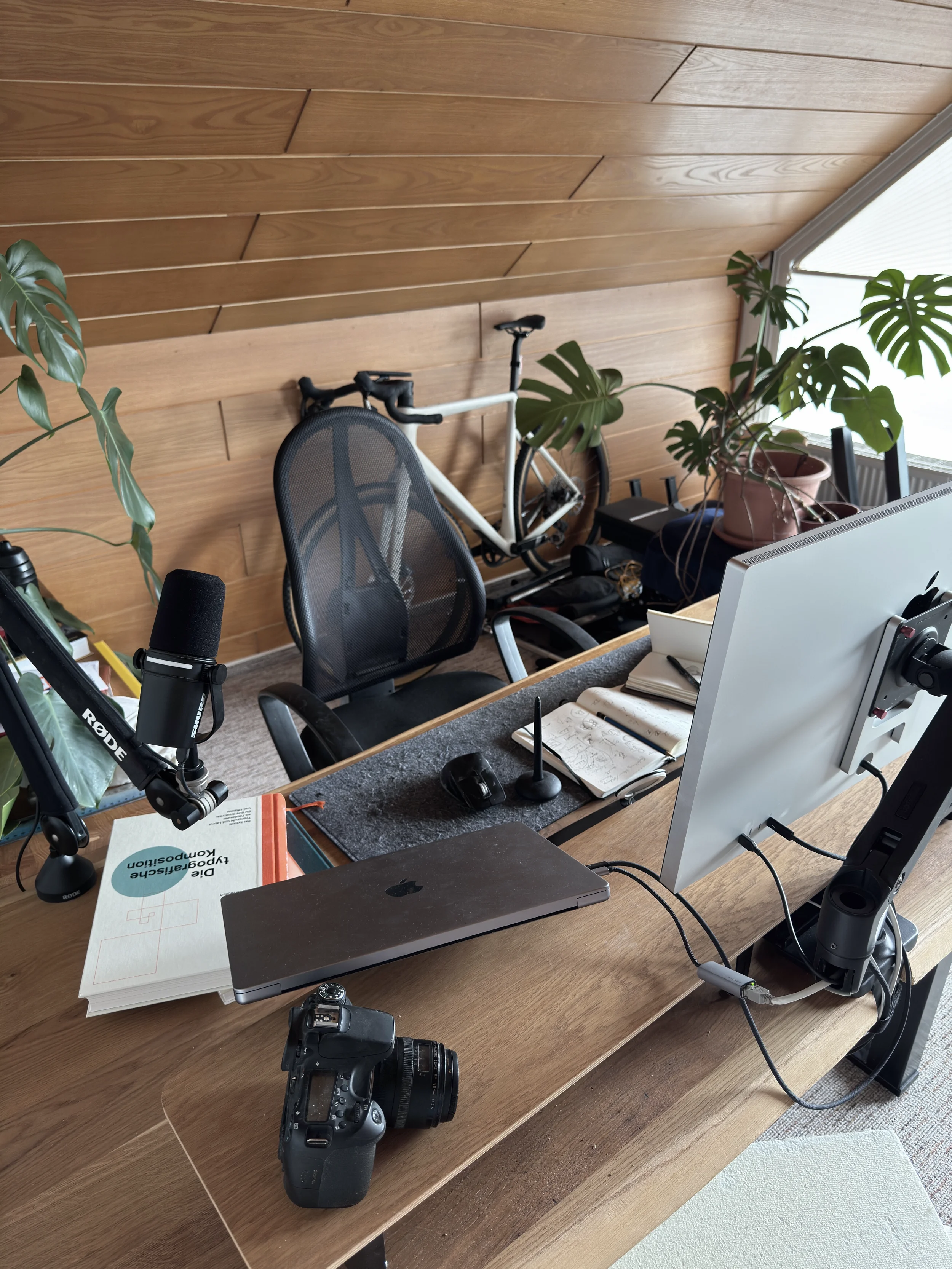 Kreativer Arbeitsplatz von FUXSTUDIO mit Holzschreibtisch, Monstera-Pflanze, Grafiktablett, Monitor, Kamera, Mikrofon, Büchern und Designwerkzeugen.
