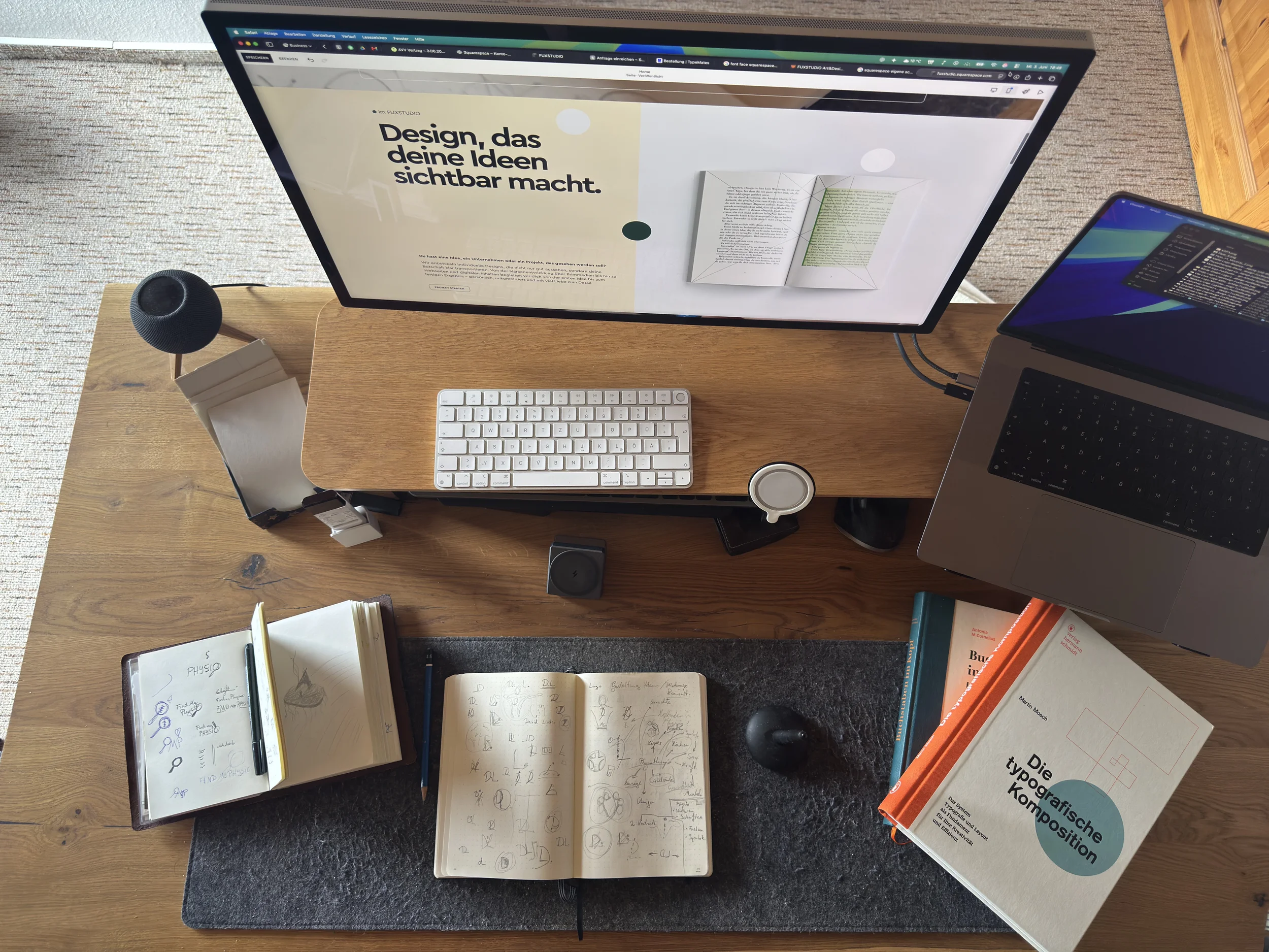Ein Schreibtisch mit einem großen Computerbildschirm, einer Tastatur, einer Tasse, einem Notizbuch mit Skizzen und Notizen, einem Laptop, mehreren offenen Büchern und einem Notizblock.