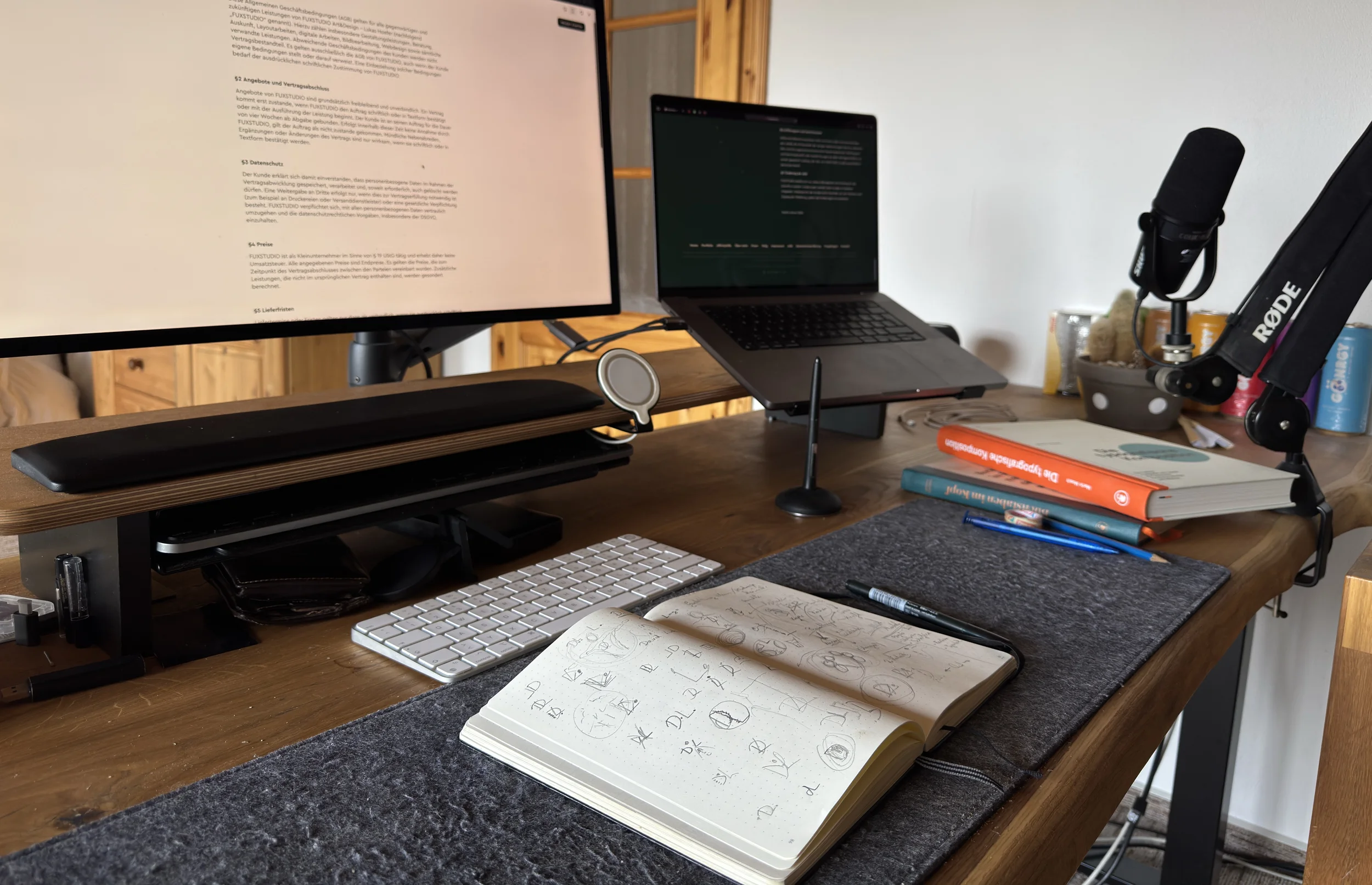 Schreibtisch bei FUXSTUDIO mit aufgeschlagenen Notizbüchern und Skizzen, im Hintergrund Bücher über Typografie und Kakteen. Links ein Monitor und ein aufgeklappter Laptop.