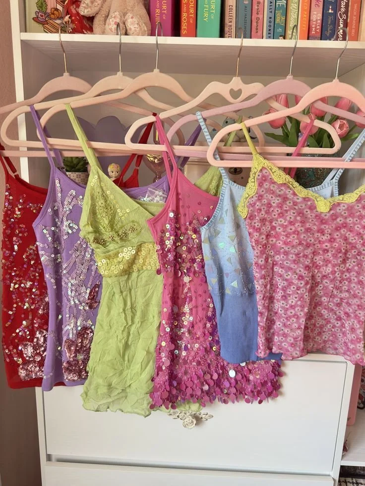 Colorful dresses hanging on pink and purple hangers on a clothing rack, with a background of books and a potted plant.