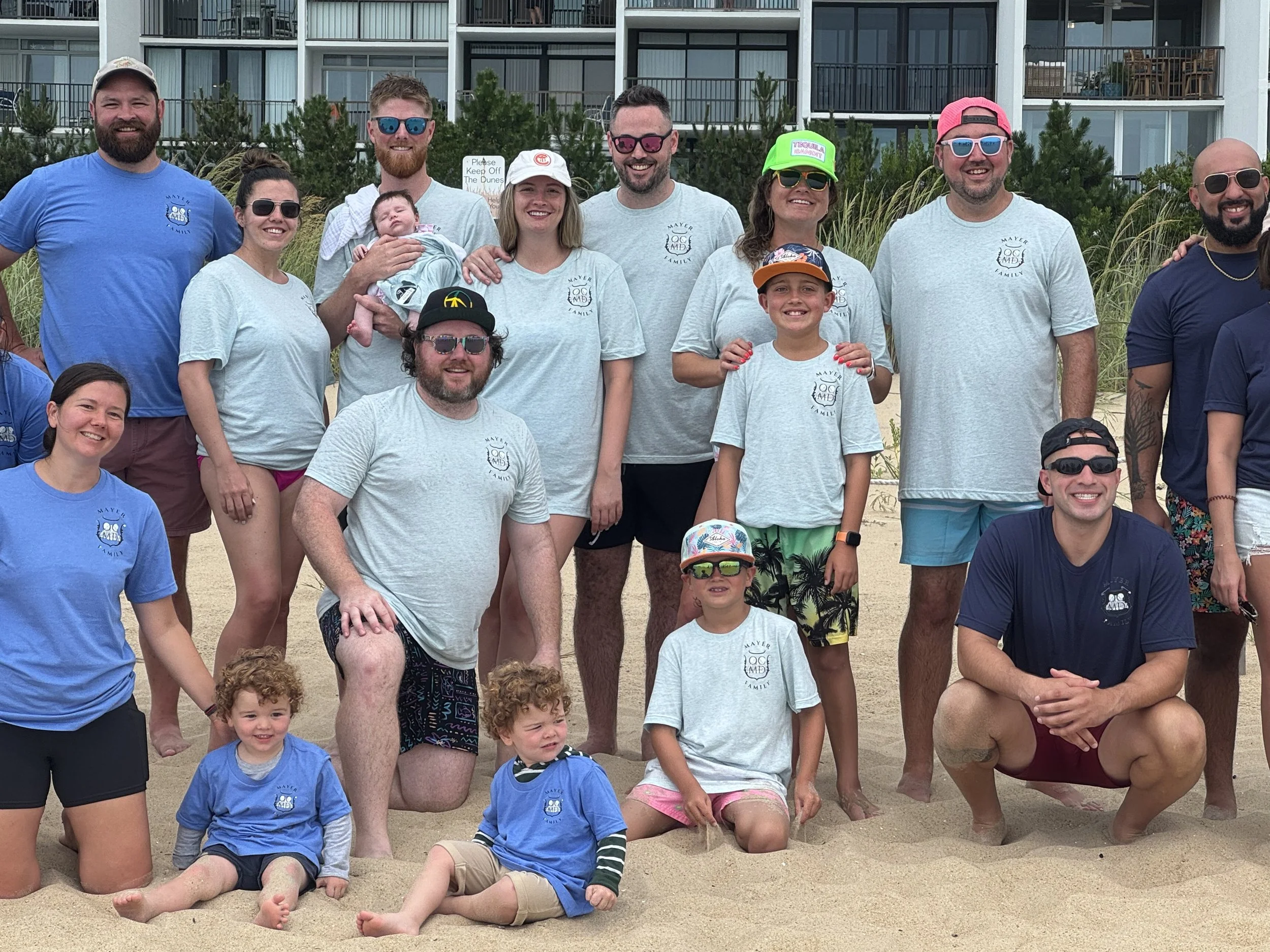 A group of happy people, including children and adults, posing on a sandy beach with apartment buildings and greenery in the background. Some are wearing matching light gray T-shirts with a logo, and others are in casual summer clothing and sunglasse