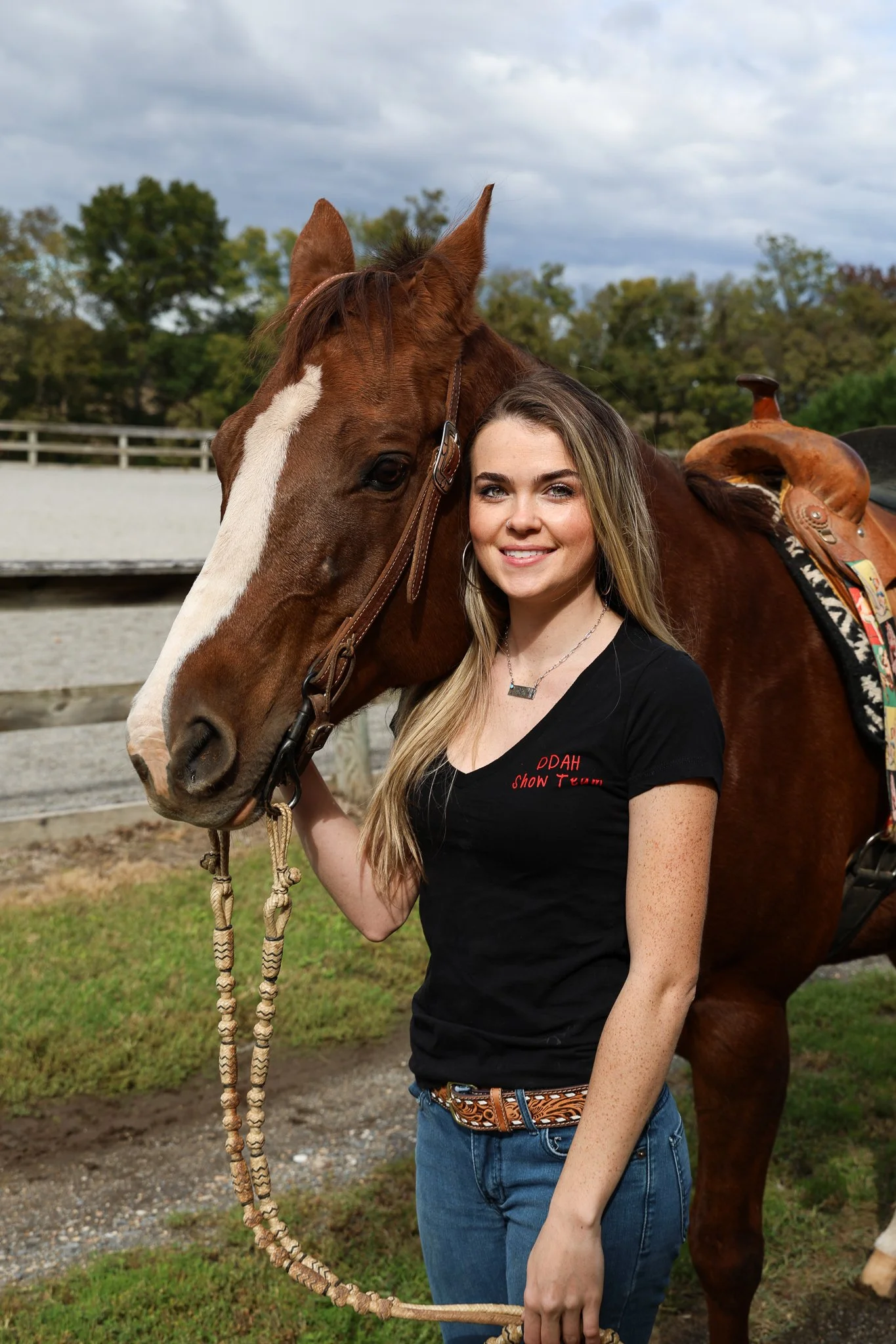 A woman with long blond hair, wearing a black t-shirt and jeans, standing next to a brown horse with a white stripe on its face, outdoors with trees and a cloudy sky in the background.