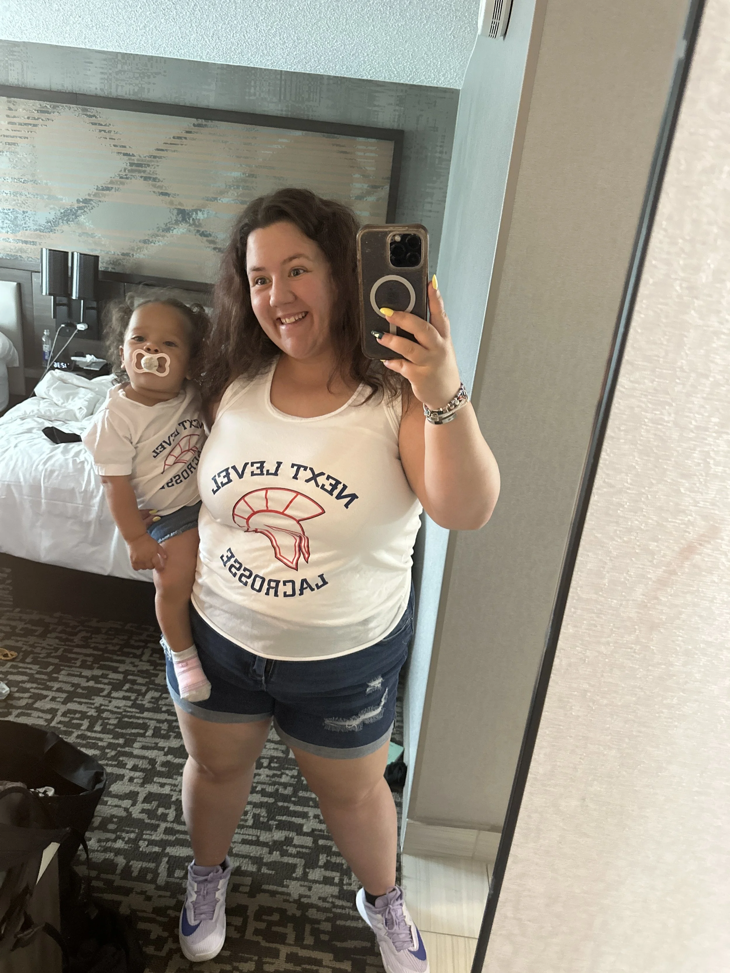 A woman taking a selfie in a hotel room mirror with a young girl on her back. The woman has curly brown hair, is wearing a white tank top and denim shorts. The girl has a pacifier in her mouth, curly hair, and is wearing a white shirt and shorts. The