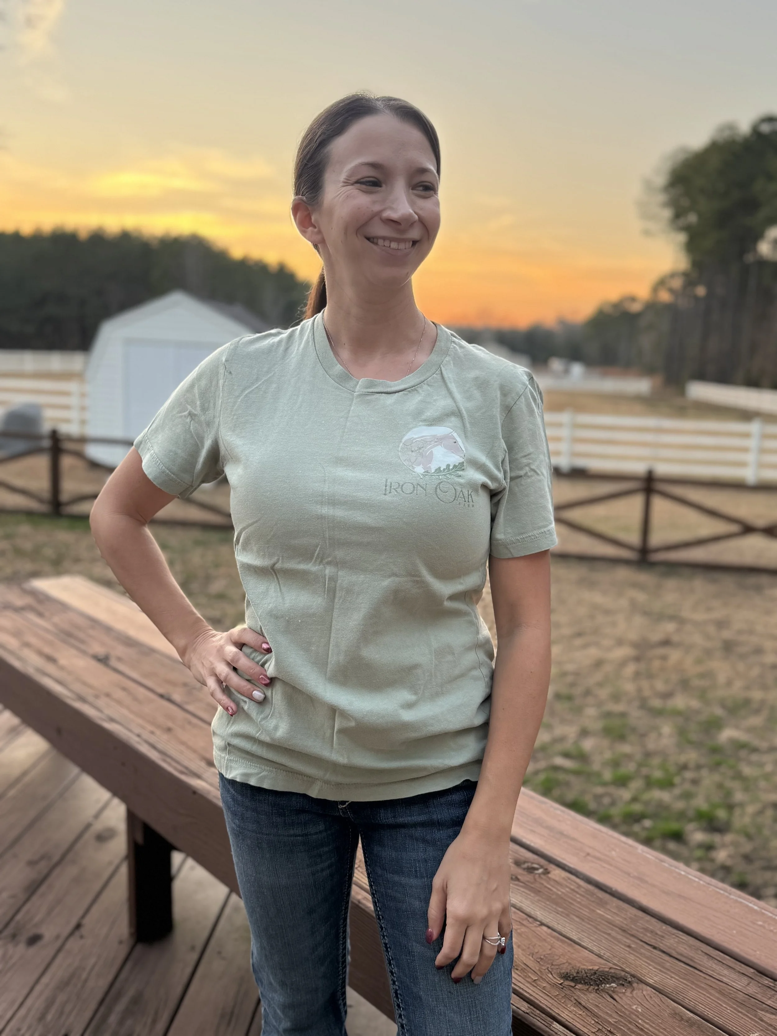 A woman with brown hair tied back, wearing a light green "Iron Oak Farm" t-shirt and blue jeans, standing outdoors on a wooden deck during sunset, with trees and a white fence in the background.