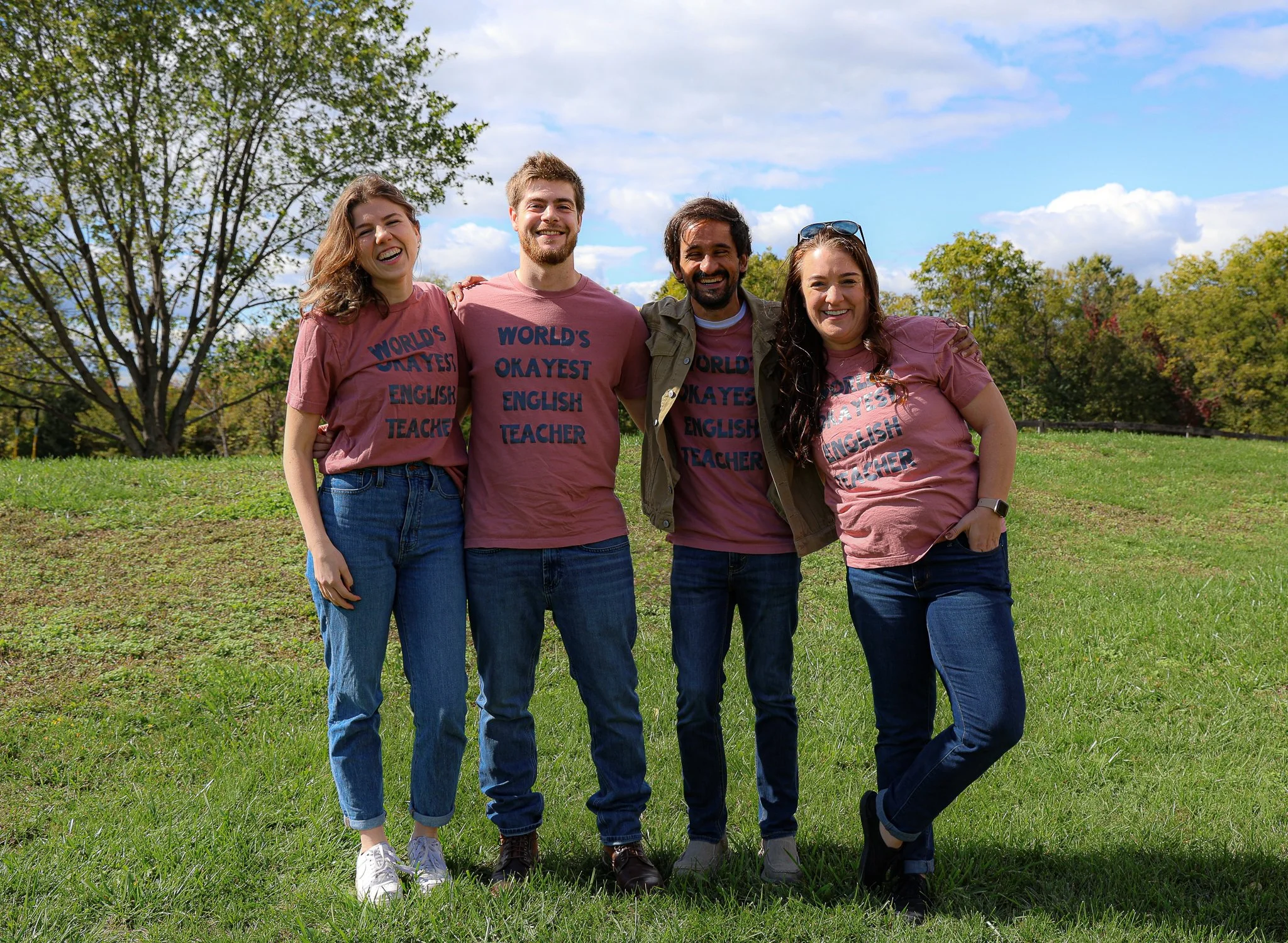 Four people standing close together outdoors, smiling, in a park with trees and blue sky in the background. They are wearing matching pink T-shirts that say 'World's Okayest English Teacher.'