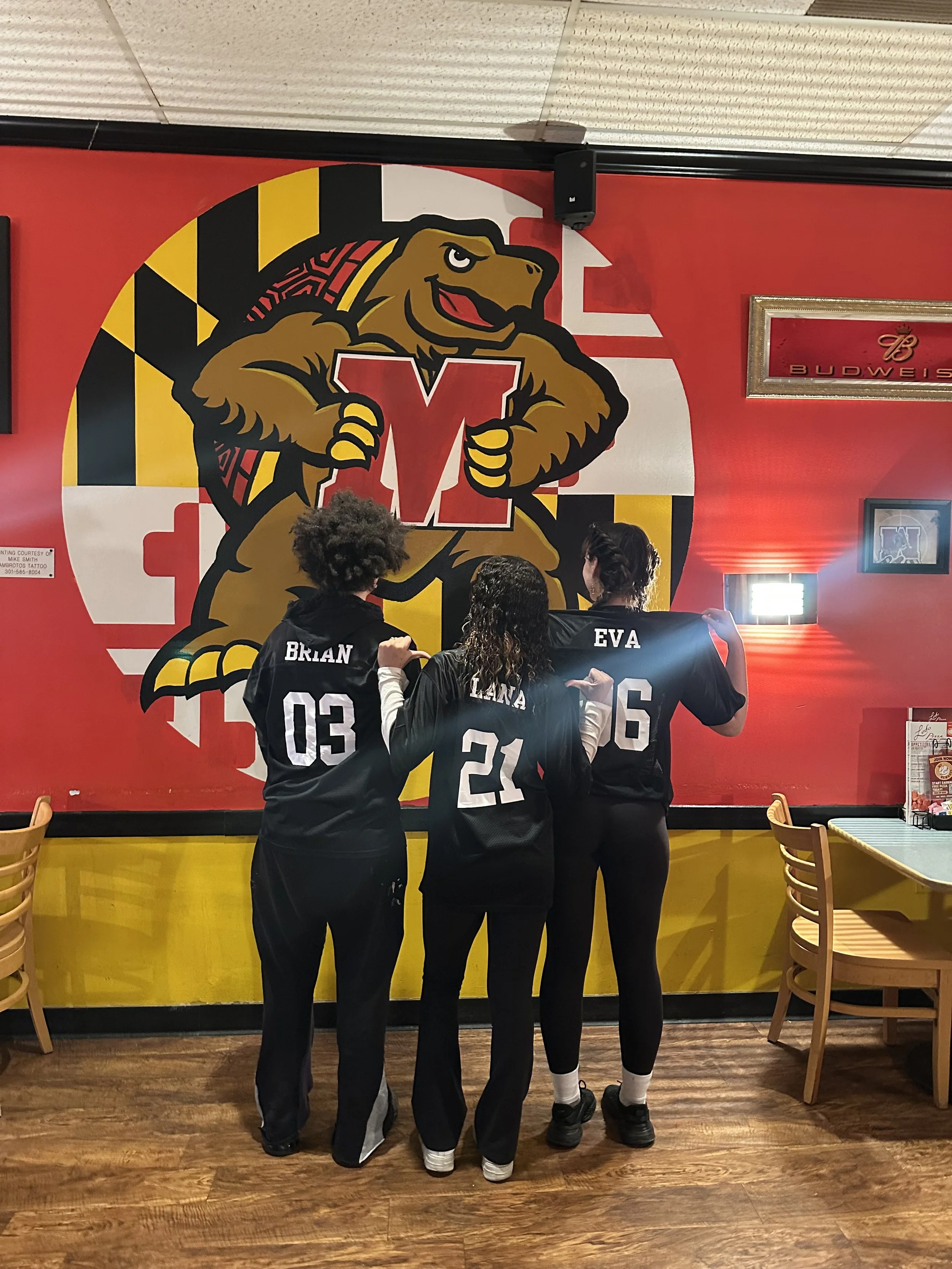 Three women wearing black basketball jerseys with white numbers and names on the back, standing in front of a large Washington Wizards logo mural inside a restaurant or sports bar, with wooden chairs and tables visible.