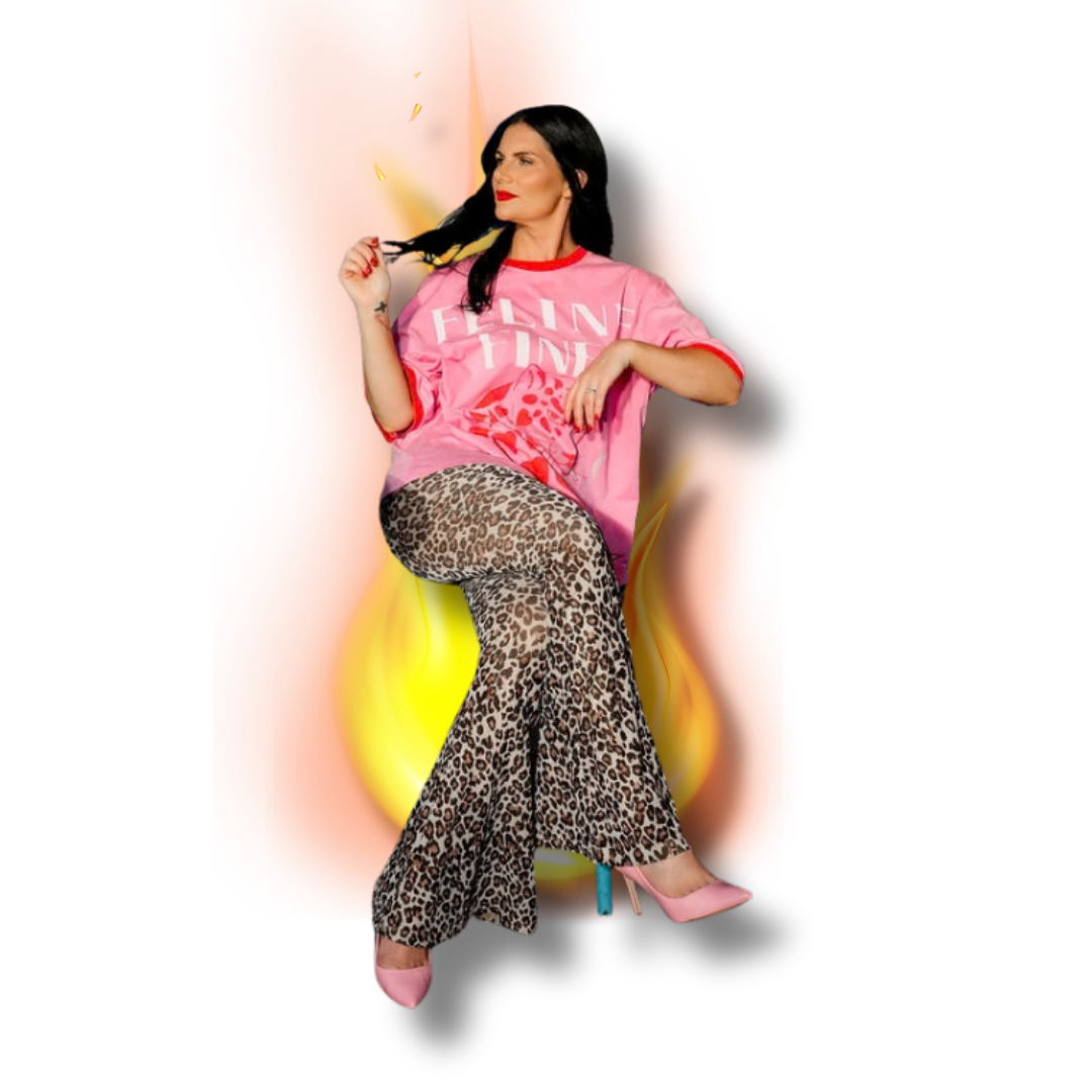 A woman with black hair and red lipstick wearing a pink t-shirt with red accents, leopard print pants, and pink high heels, seated against a fiery background.