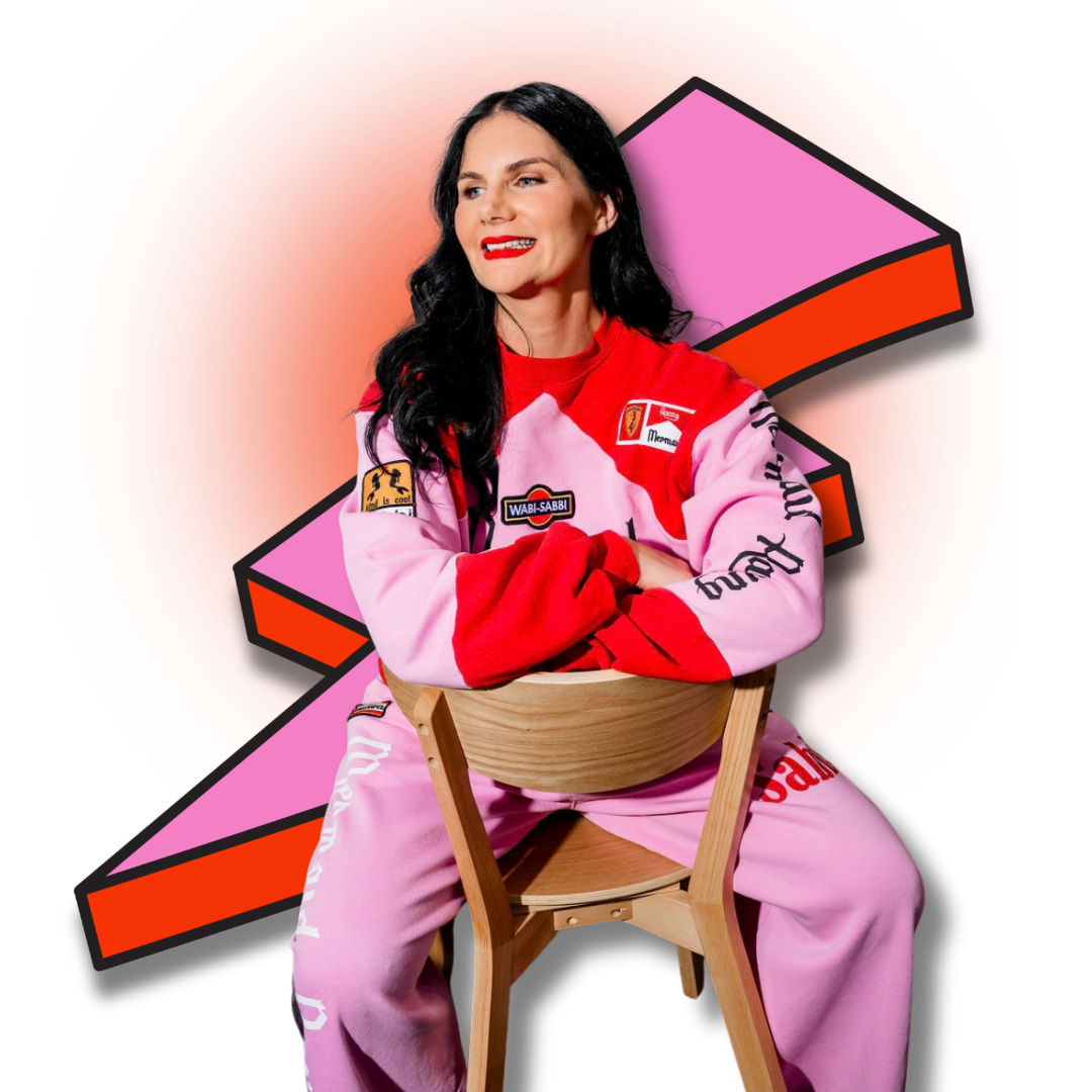 A woman with black hair and red lipstick, wearing a pink and red racing jacket with various patches, sits on a wooden chair with her arms crossed, smiling. The background features a cartoon-style pink slice of cake with red and black outlines on a bright red backdrop.