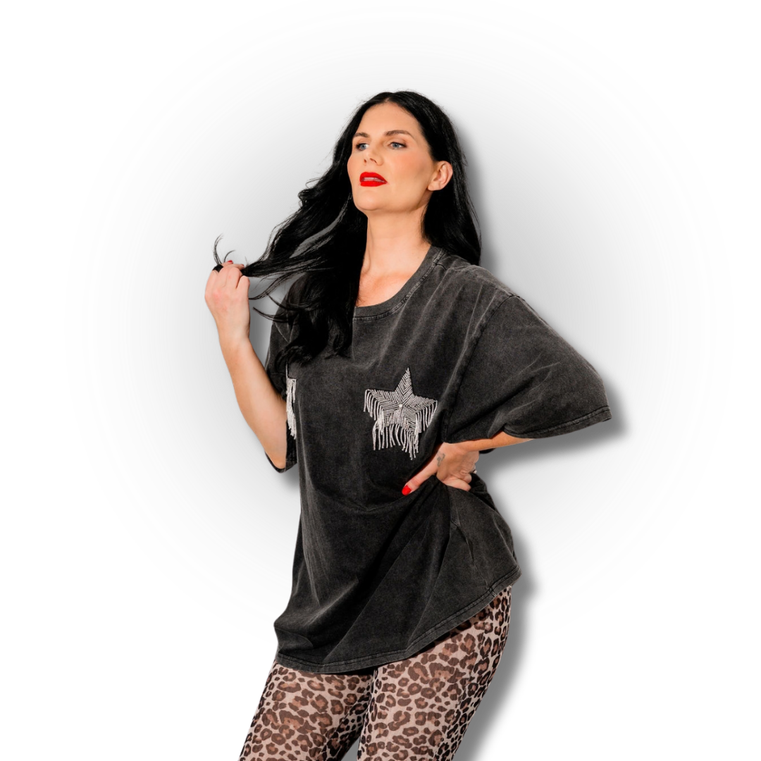 A woman with long black hair, wearing a loose dark grey T-shirt with a star design, and leopard print pants, posing with one hand on her hip and the other holding her hair, against a circular gradient background.
