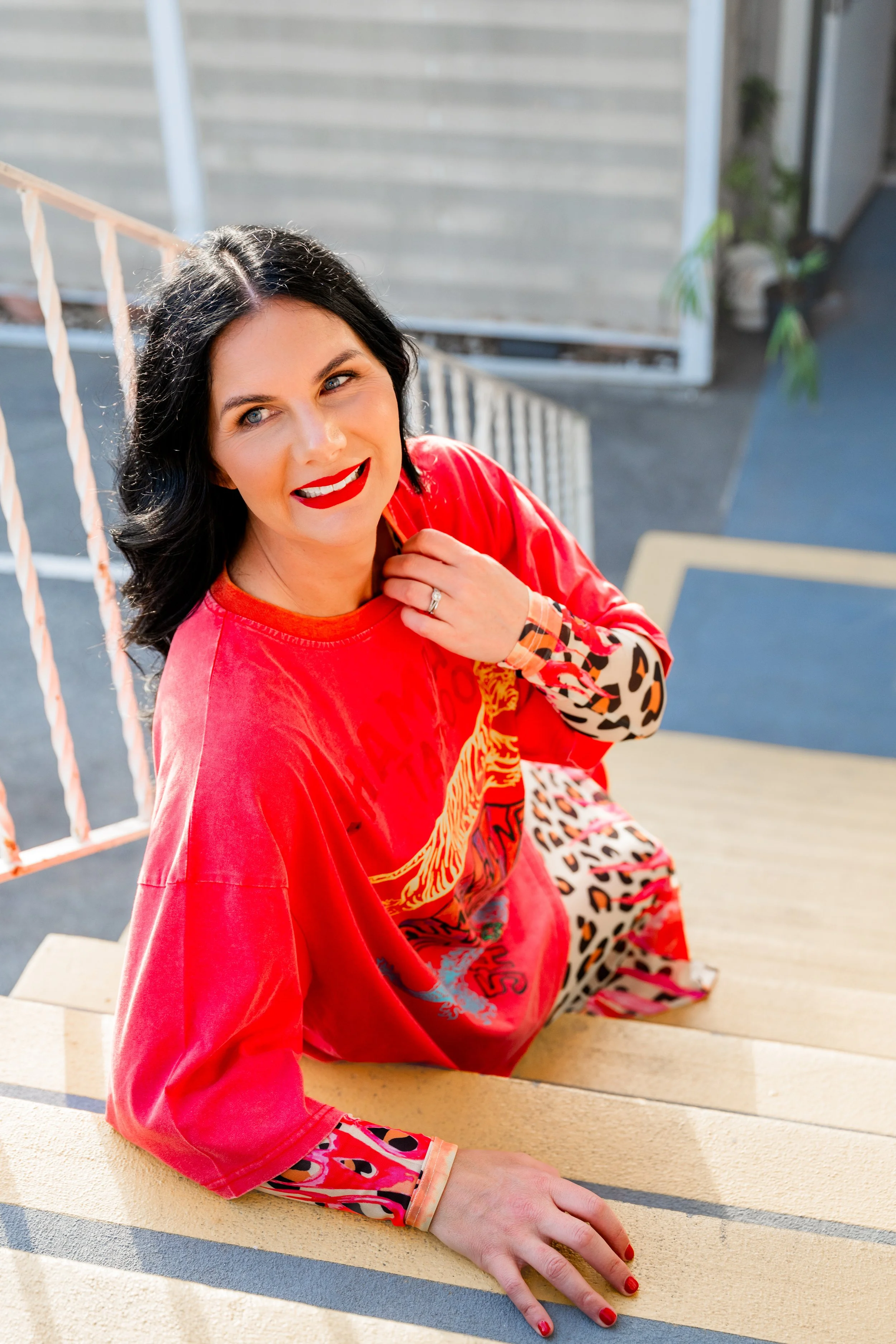 A woman with black hair and red lipstick sitting on outdoor steps, wearing a red shirt with animal print sleeves and pants with a colorful animal print pattern, looking up and smiling.