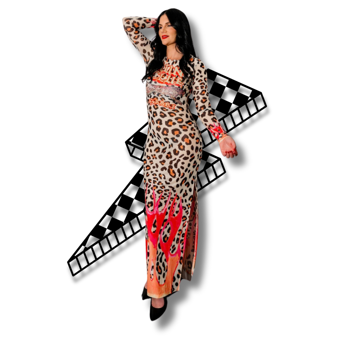 Woman with long black hair wearing a leopard print dress with red and pink flame designs, posing with one hand on her head and the other by her side, against black background.