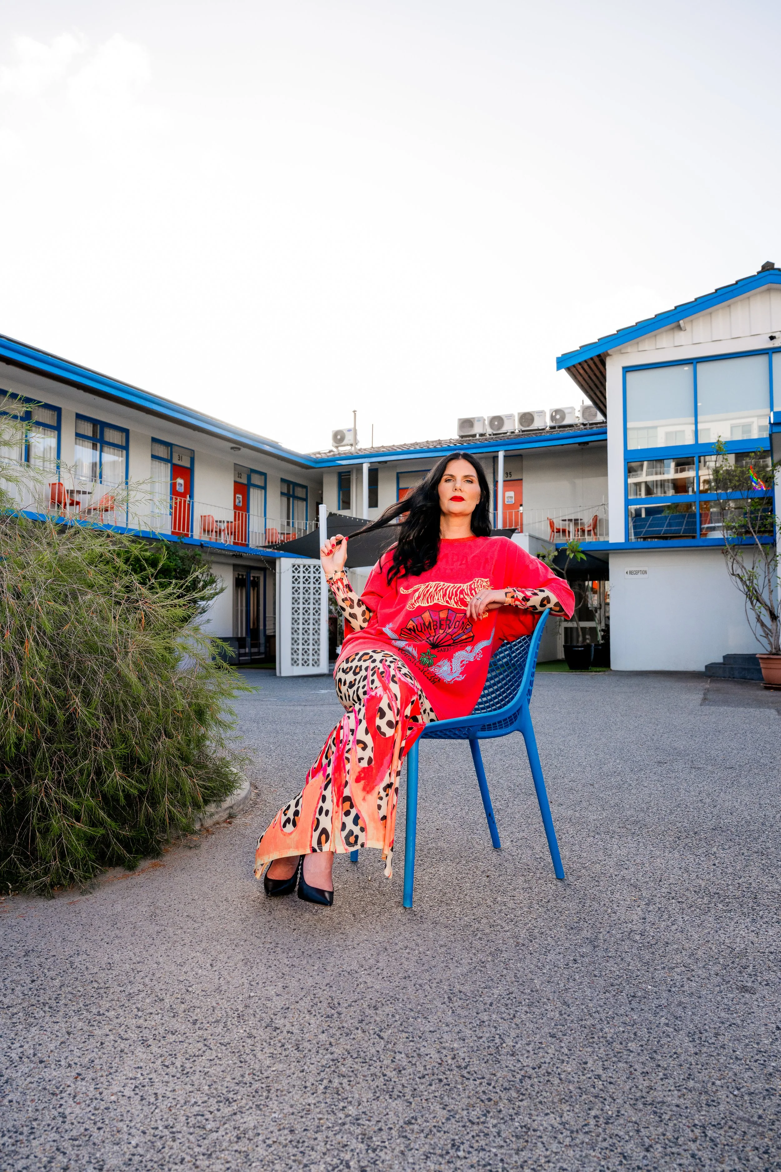 A woman with long dark hair sitting on a blue plastic chair outside a motel or apartment complex. She is wearing a red T-shirt with a colorful graphic, leopard print pants, and high heels. She has her hand in her hair and is looking directly at the camera. The background features a building with blue accents, multiple doors, and outdoor air conditioning units.