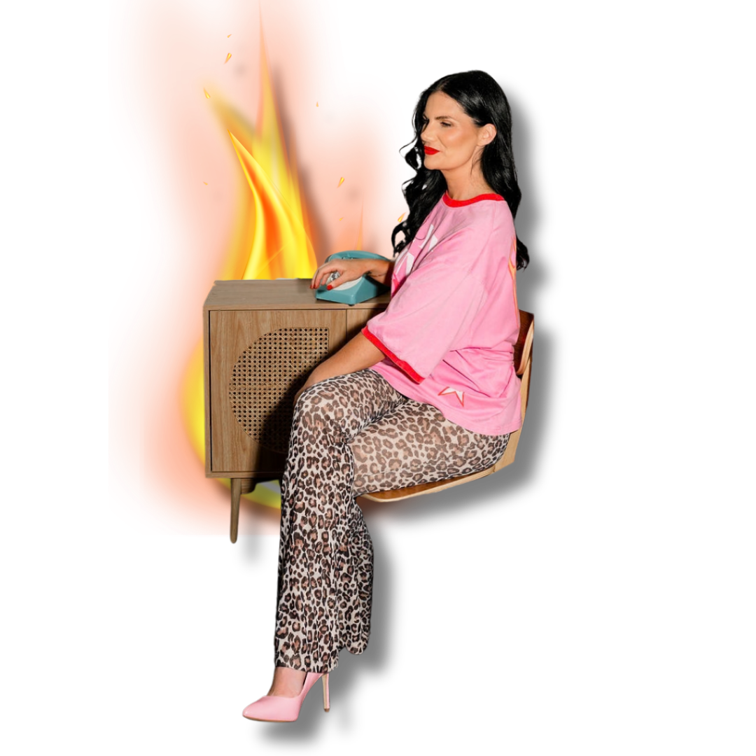 A woman with long dark hair wearing leopard print pants, pink high heels, and a pink shirt with red accents, sitting on a chair next to a vintage wooden radio. She is operating a teal rotary phone. The background is edited with a large graphic of flames and a black backdrop.
