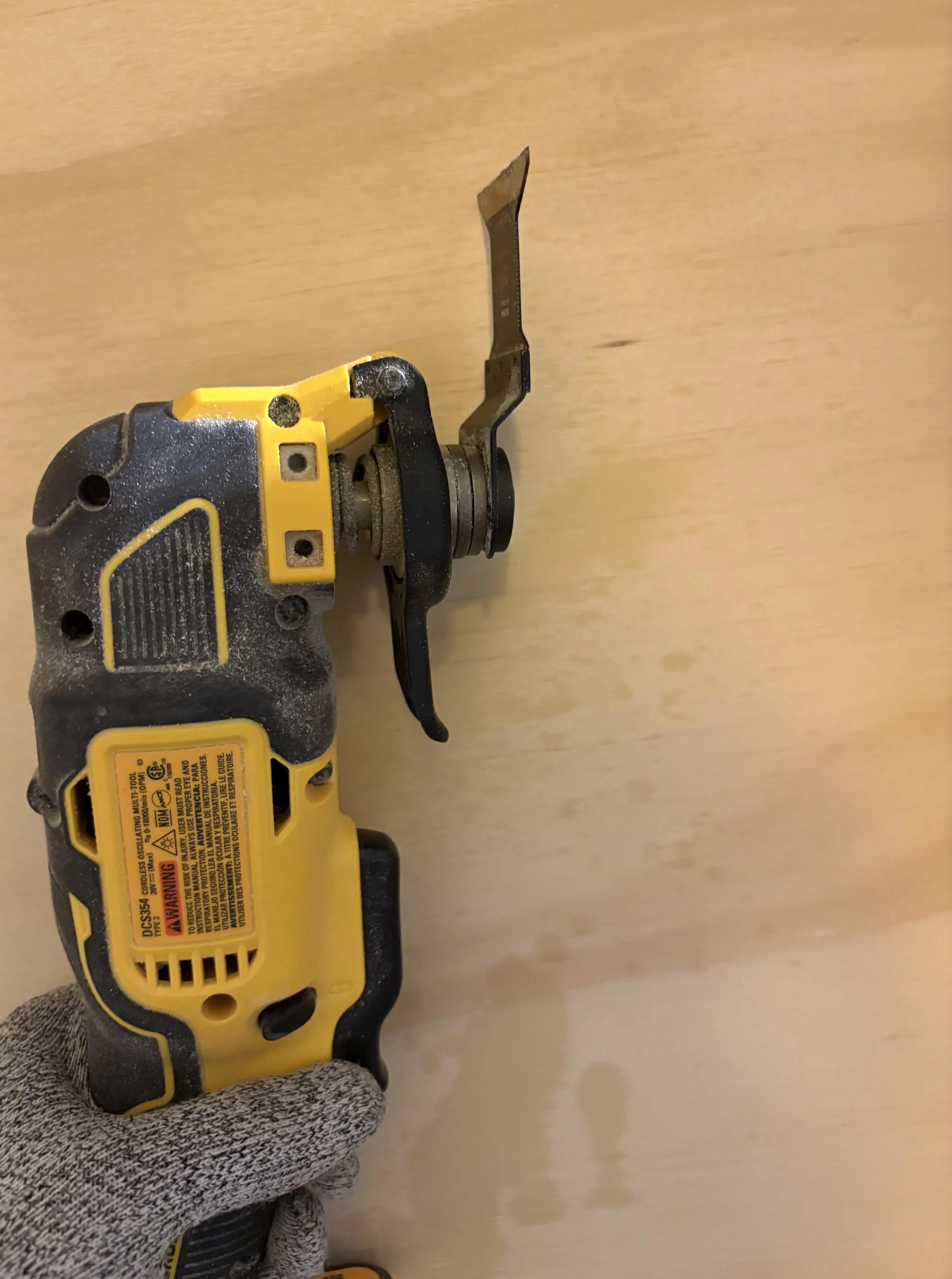 A person wearing a gray glove holding a yellow and black cordless power tool, possibly a multi-tool, with a blade attachment, against a light-colored wooden surface.