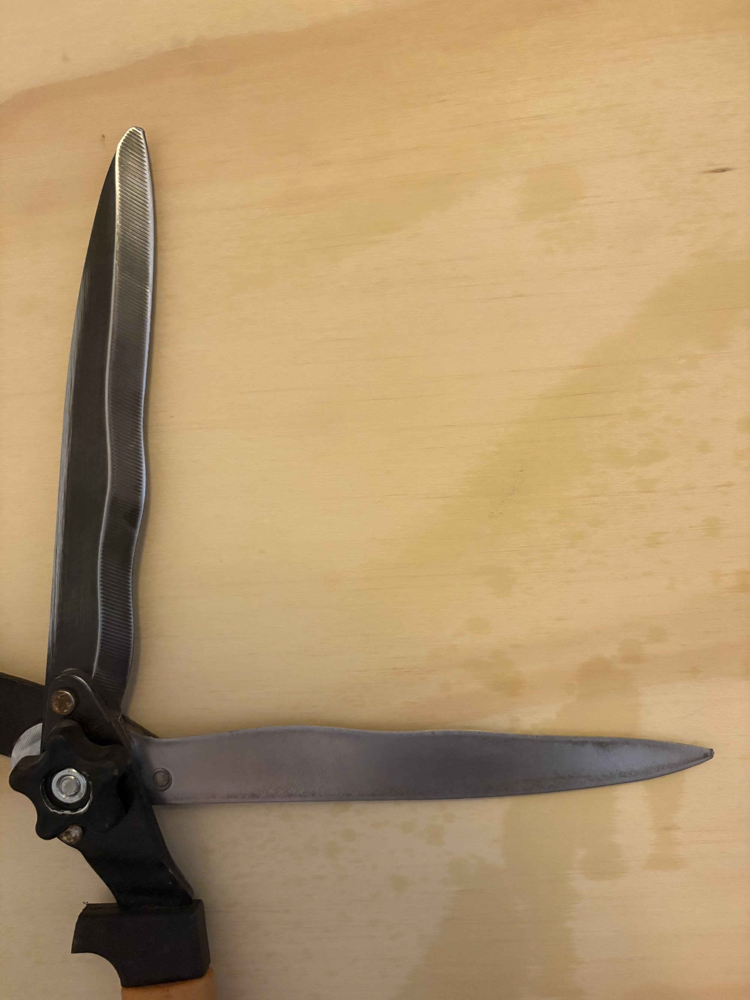 A knife with a black handle and a wide, flat, dark blade resting on a light wooden surface.