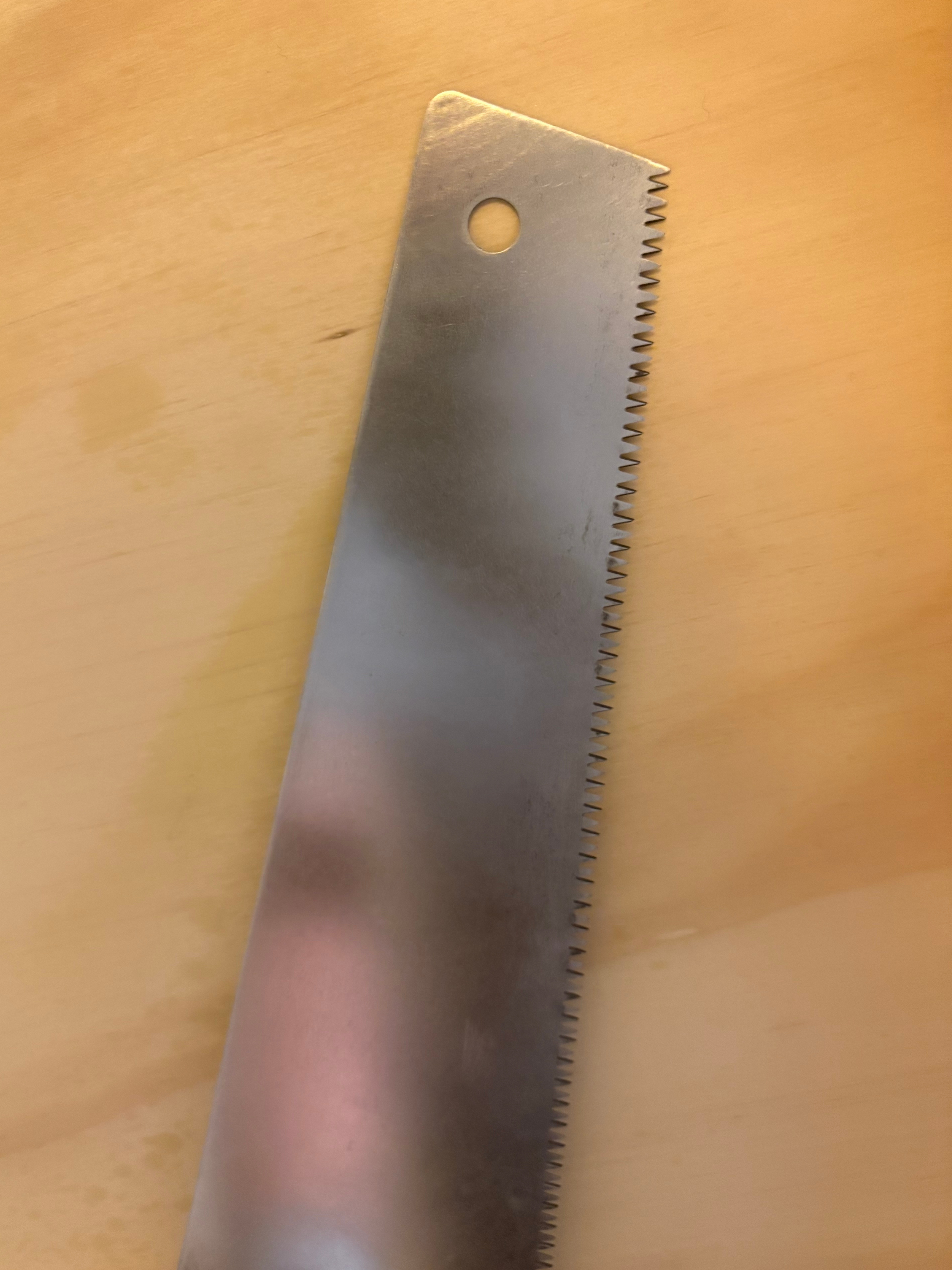 A metal saw blade with jagged teeth and a circular hole near the top, placed on a light-colored wooden surface.