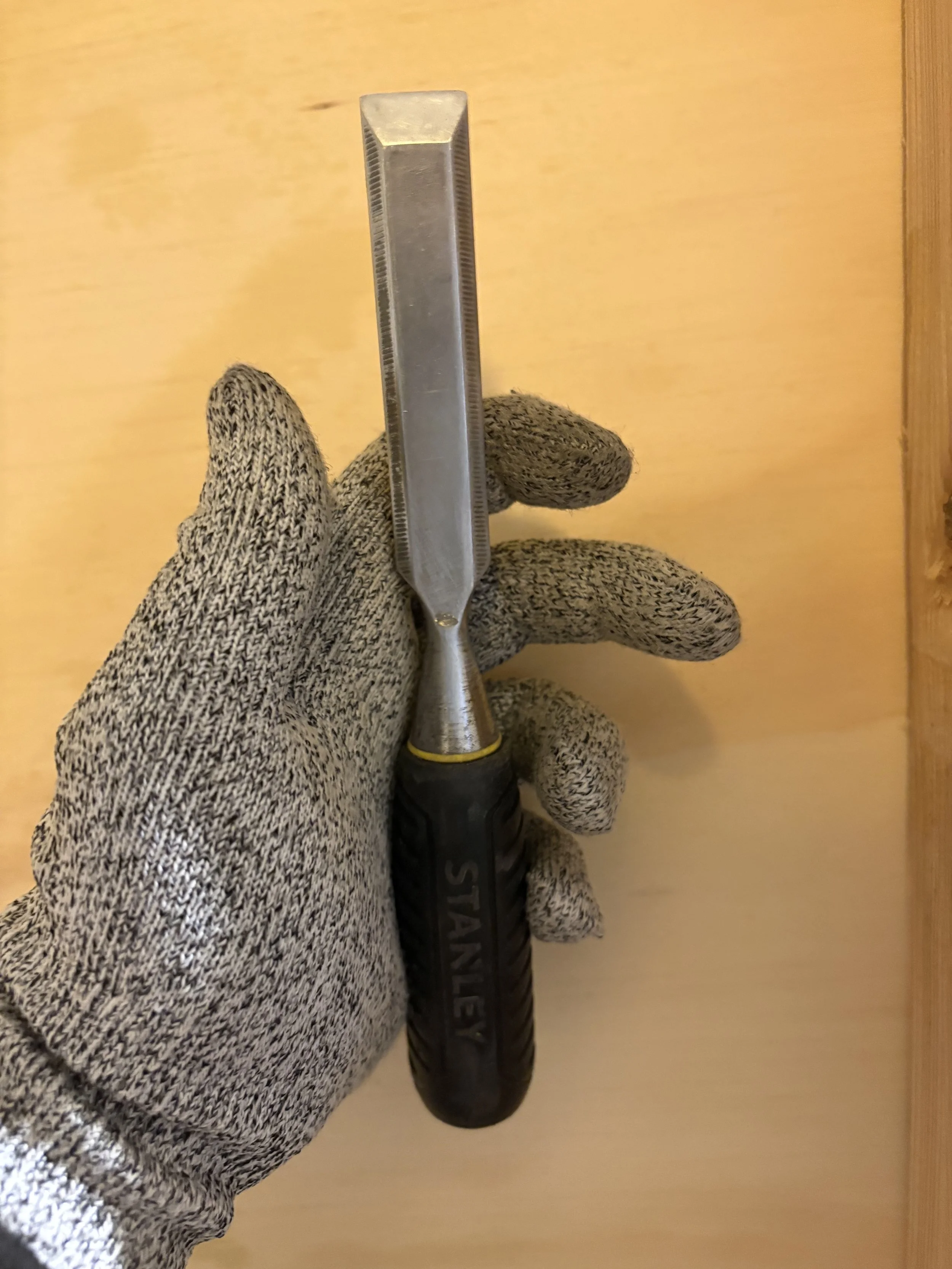 Close-up of a gloved hand holding a metal nail puller tool with a yellow ring near the handle, against a wooden background.