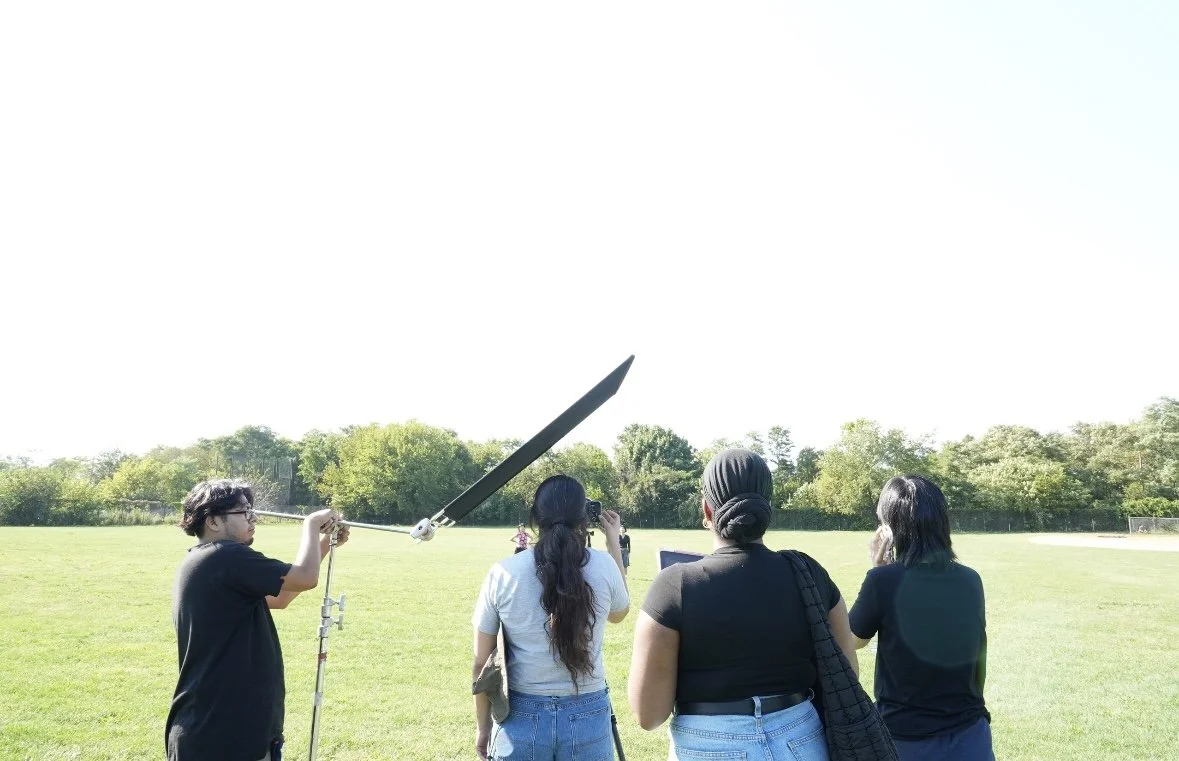 Group of four women and one young man on a grassy field, with the man holding a large sword upright in the air, and the women standing nearby, some taking photos.