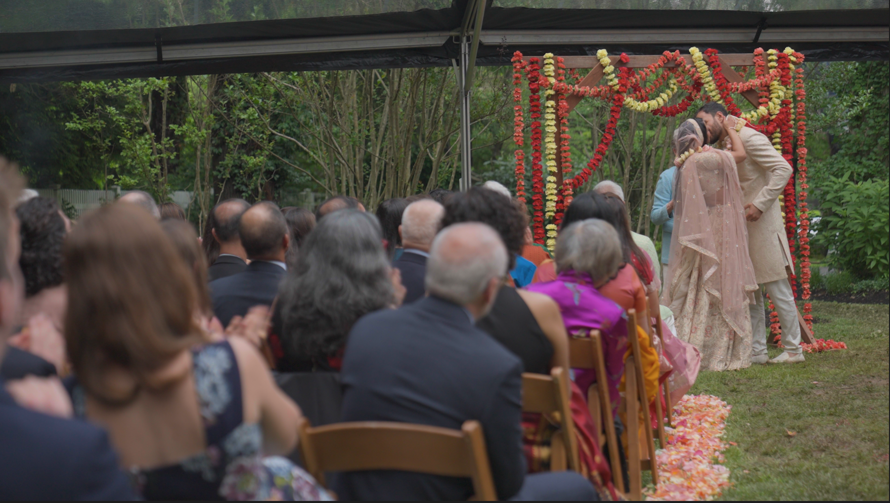 A couple wearing traditional Indian wedding attire embracing each other during an outdoor wedding ceremony, with guests seated and watching in the background.