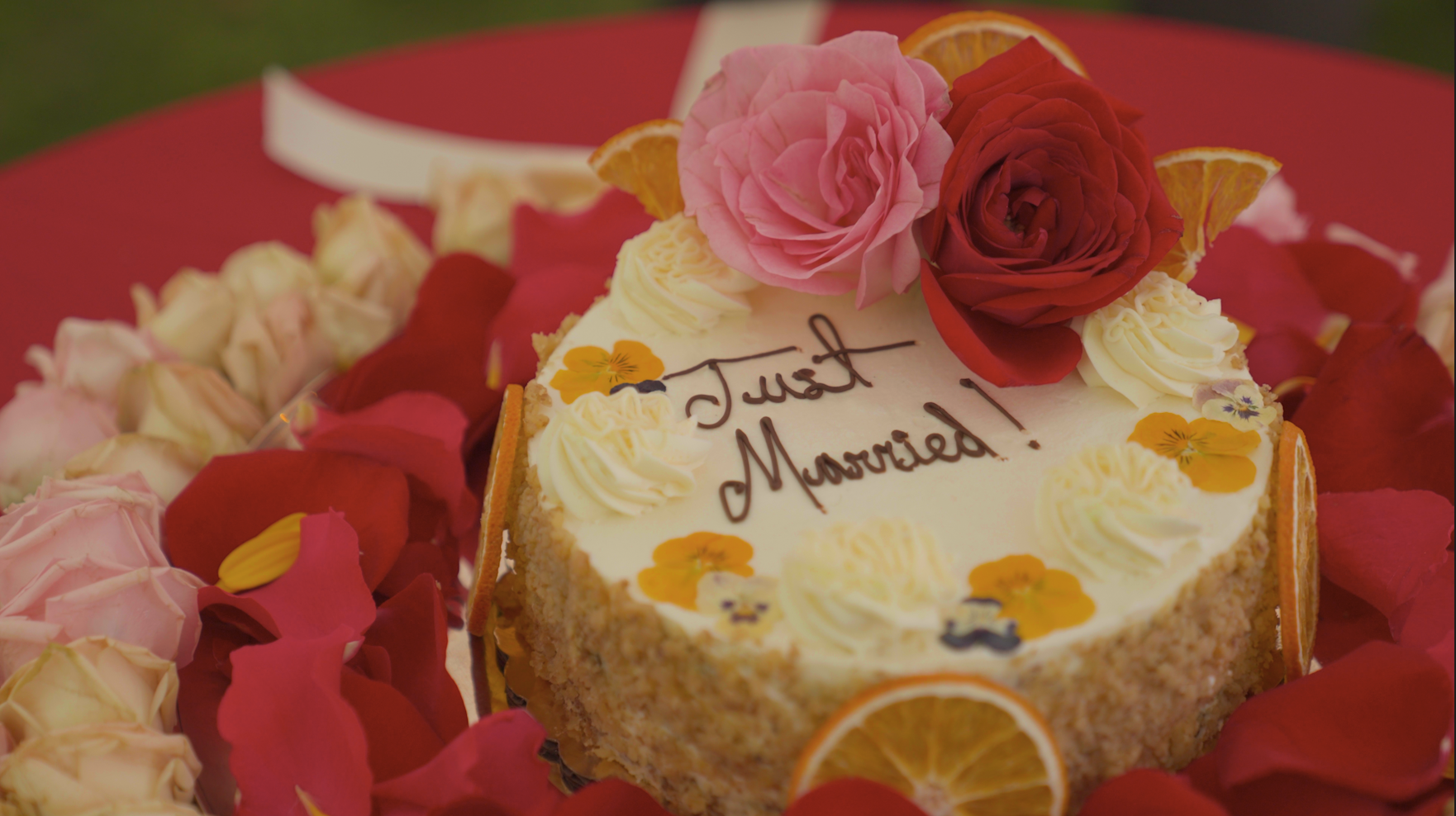 A round celebration cake decorated with pink, red, and white roses, orange slices, and edible flowers, with the message "Just Married!" written in chocolate on top, surrounded by pink, red, and cream-colored flower petals.