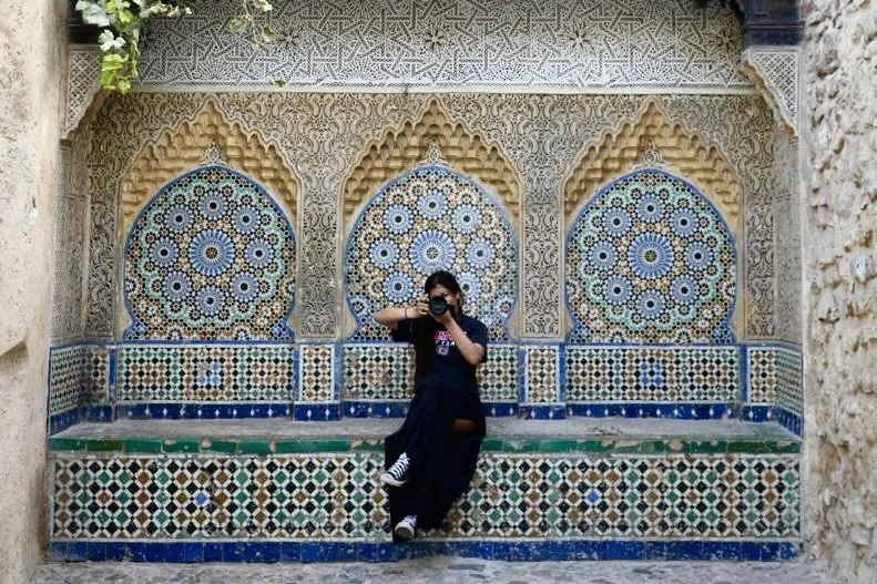 Person sitting on a decorative stone and tile bench in front of an intricate Moroccan-style mosaic wall with arched niches.