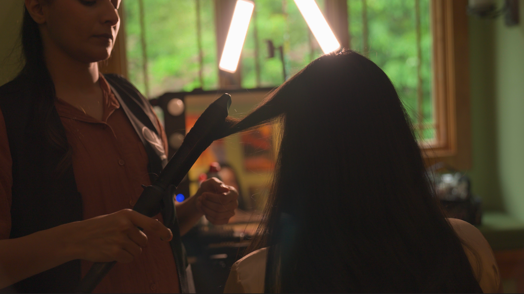 A hairstylist curling a woman's long dark hair in a salon with large windows showing green trees outside.