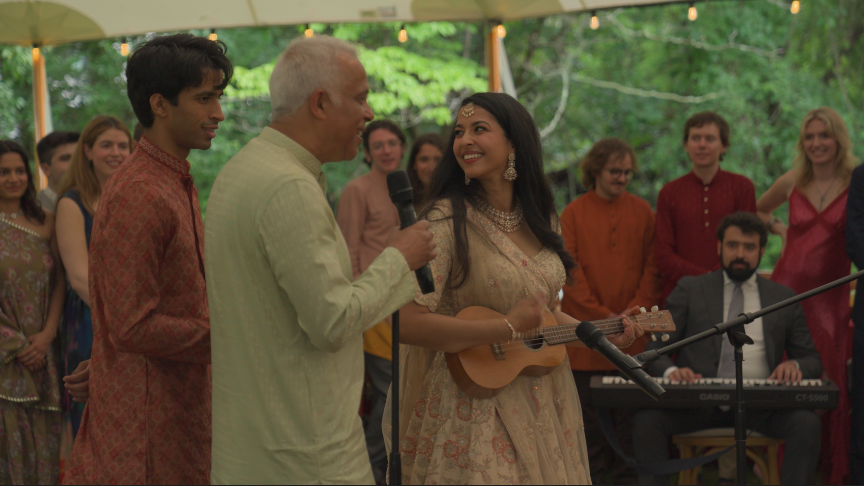A group of people performing at a celebration outdoors, with two individuals singing or speaking into microphones and a woman playing a small guitar. Others in traditional Indian attire are dancing and enjoying the event.