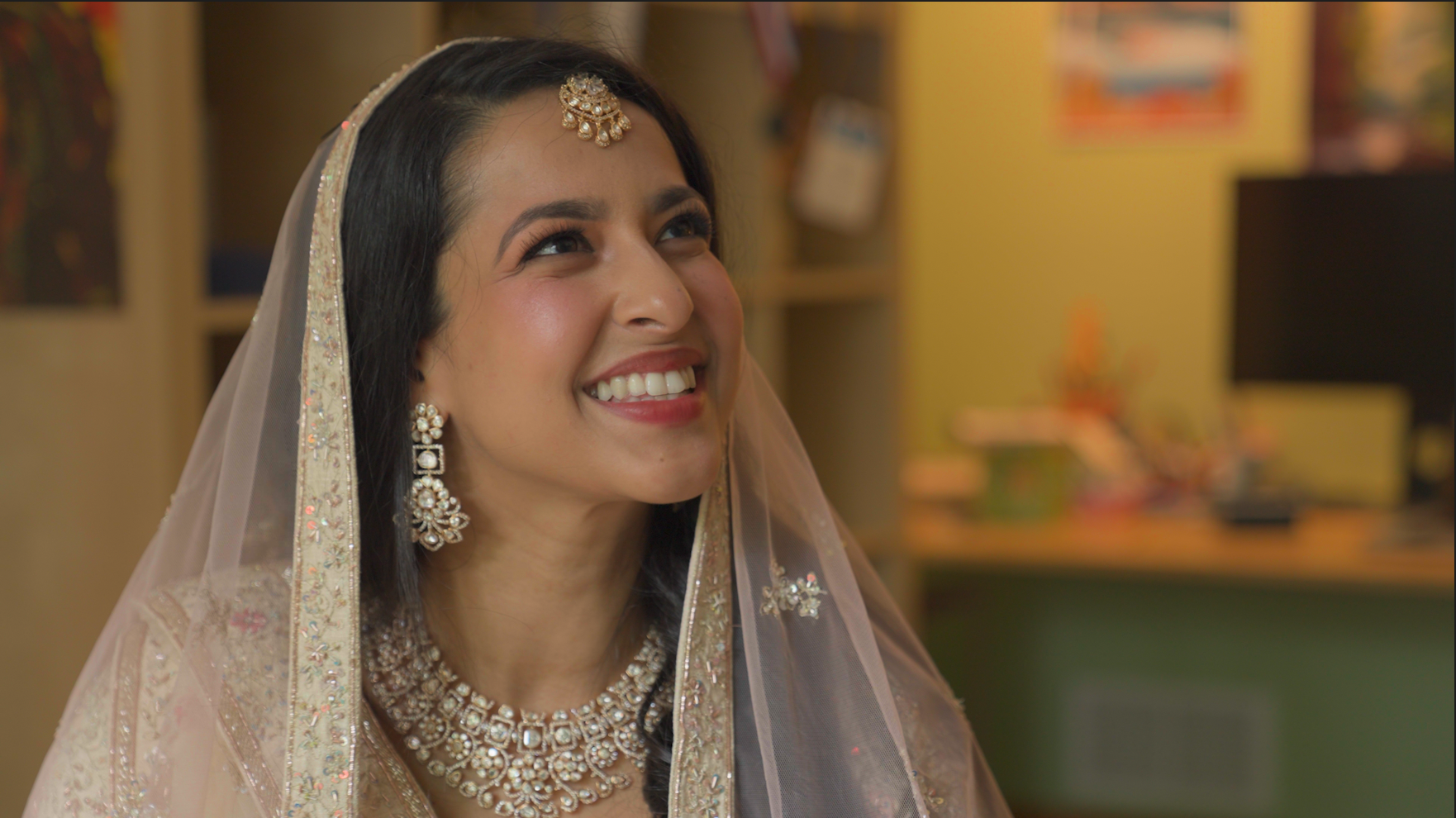 A woman dressed in traditional Indian wedding attire, wearing jewelry and a veil, smiling with a joyful expression.