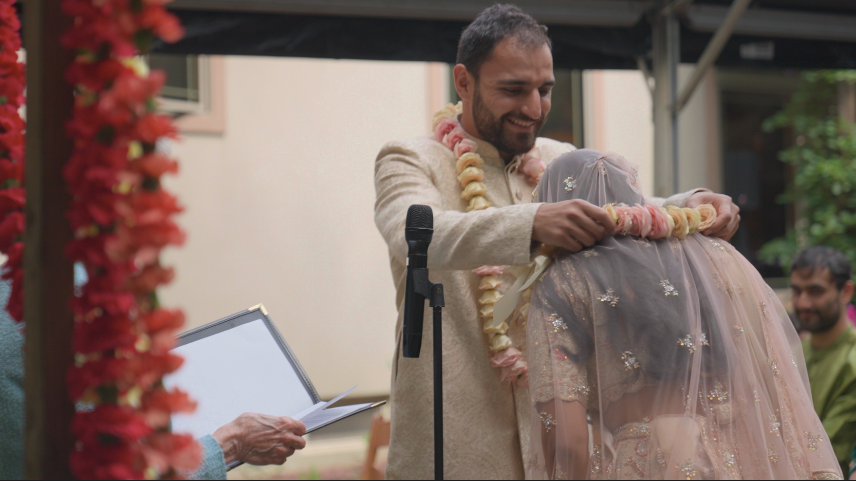 A man in traditional attire placing a flower garland on a woman dressed in a pastel pink and gold embroidered gown, during a ceremony with a microphone stand and a person holding a notepad in the background.