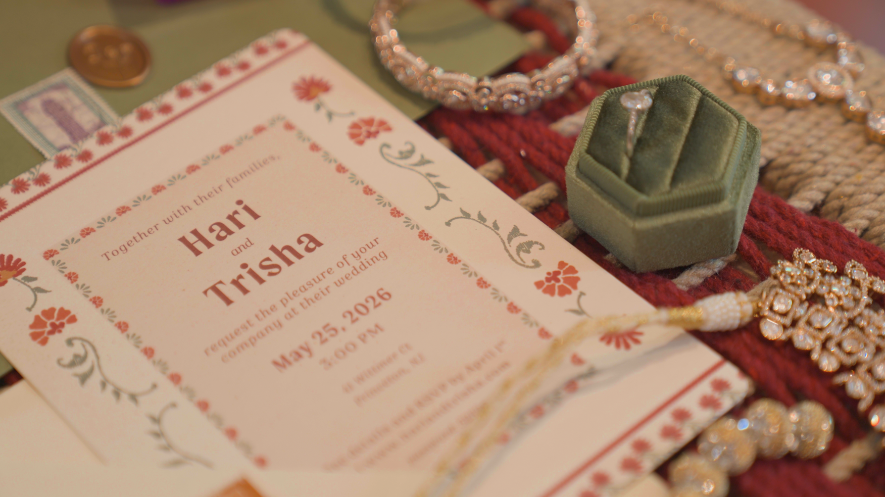 Wedding invitation with floral border, surrounded by jewelry including a diamond bracelet, a green ring box, and a red and gold decorative fabric.