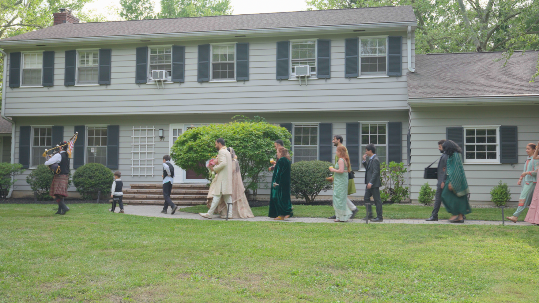 A diverse group of people walking on a sidewalk in front of a beige house with blue shutters, celebrating a wedding, with a bagpiper leading the procession.