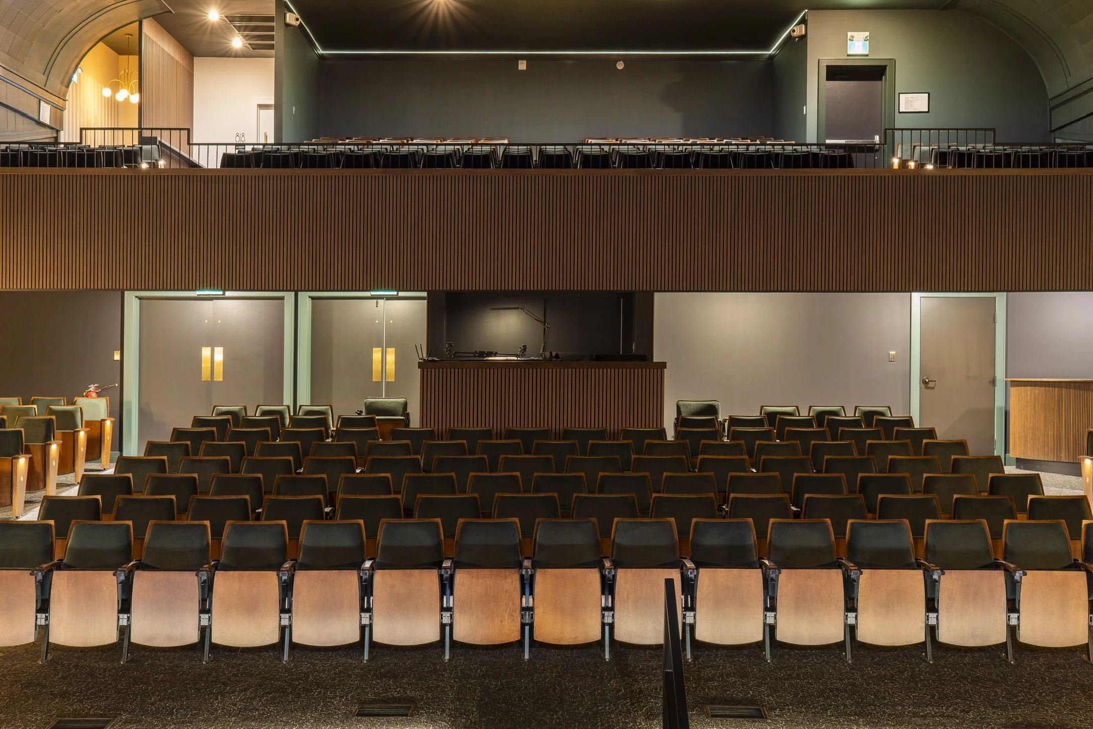 An empty tiered auditorium with rows of black and beige seats, a stage area, a dark green wall at the back, and modern lighting fixtures.