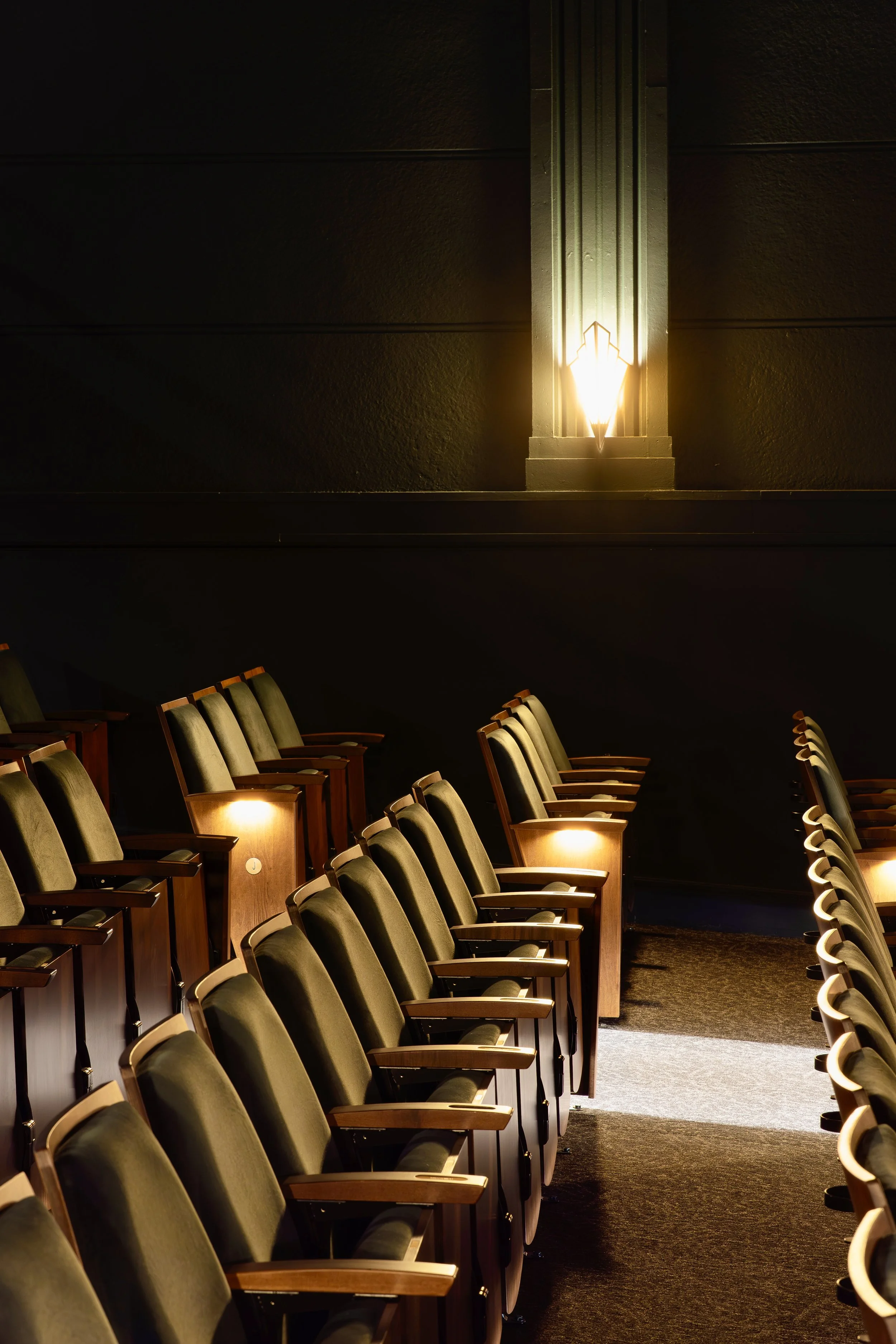 Rows of dark green theater or auditorium seats with wooden armrests, illuminated by warm lighting, with a decorative wall sconce on the wall behind.