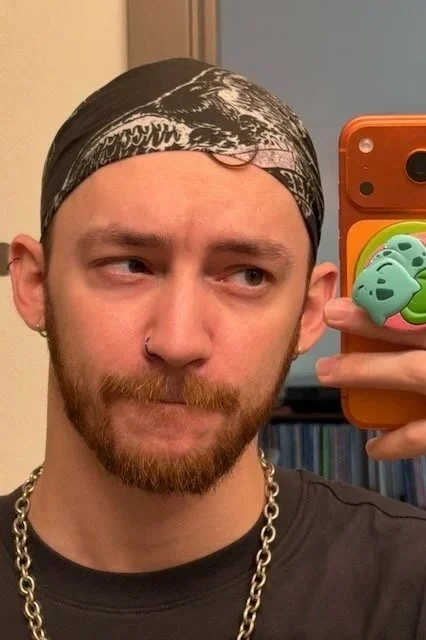 Young man with a bandana, beard, nose ring, and chain necklace taking a mirror selfie with an orange phone.