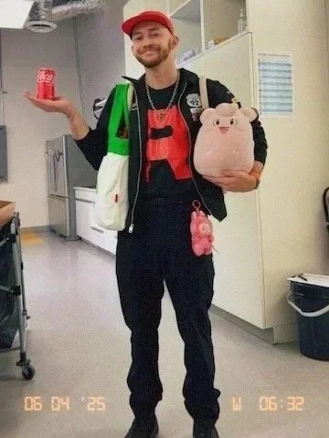 A man in a red baseball cap and black jacket, smiling and holding a can of Coke in one hand and a small plush Clefairy (Pokémon) in the other, standing in a workplace setting.