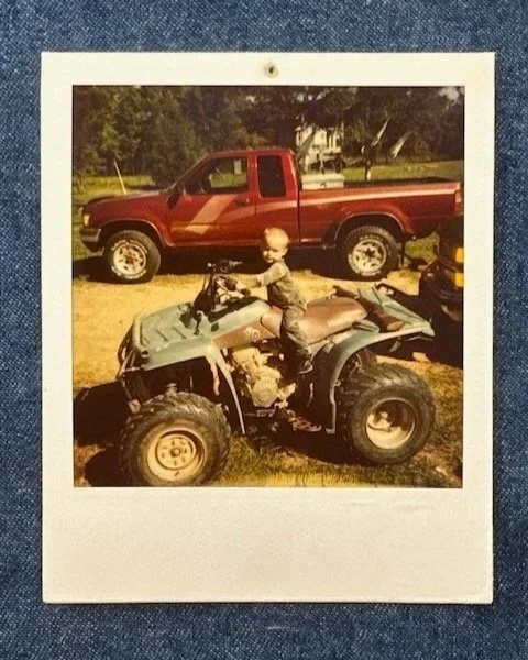 Photo of a Polaroid featuring a young boy sitting on an all-terrain vehicle (ATV) in front of a red pickup truck in an outdoor setting with trees in the background.