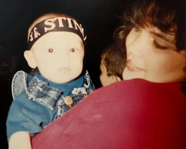 A woman is holding a young child with a headband that says 'LA STING'. The child's expression is neutral, and the woman is smiling while looking at the child.