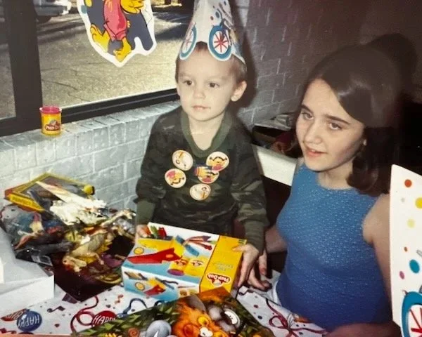 A young boy and his sister celebrating a birthday with a table full of party supplies and gifts inside a McDonald's. The boy wears a party hat with a bicycle design and cartoon buttons on his shirt, while the girl next to him wears a blue dress.