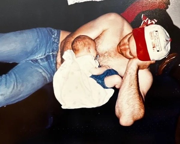 A shirtless man lying on a couch with a small child resting on his chest, both appearing to cuddle or nap together.